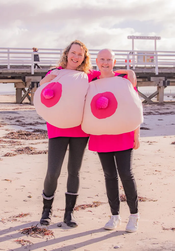 Two women standing on a beach in pink tops holding inflatable boobs