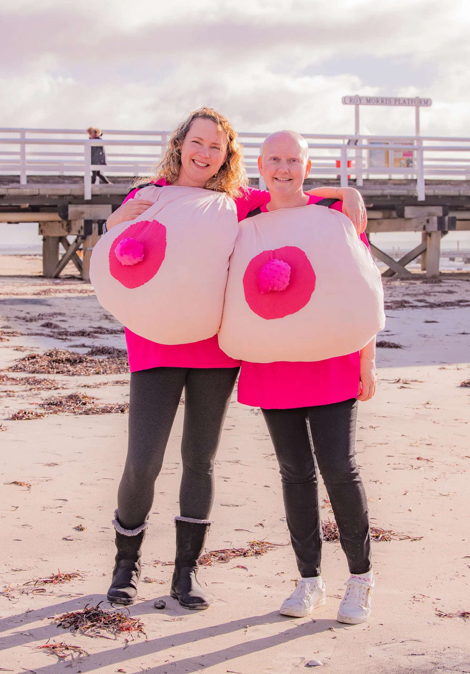 Two women standing on a beach in pink tops holding inflatable boobs 