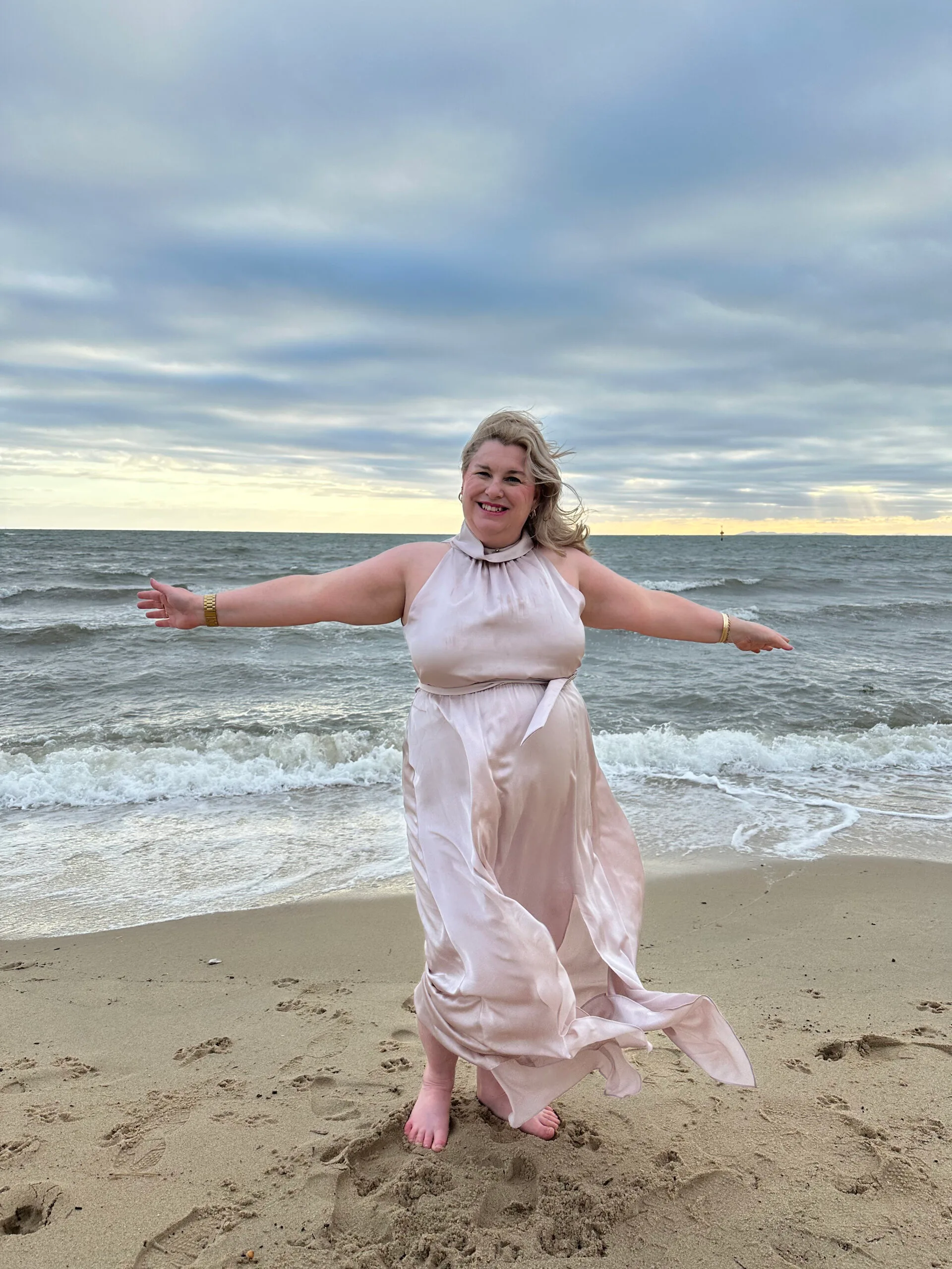 Woman with blonde hair wearing long pink gown, stretching arms and standing on beach while smiling at camera.