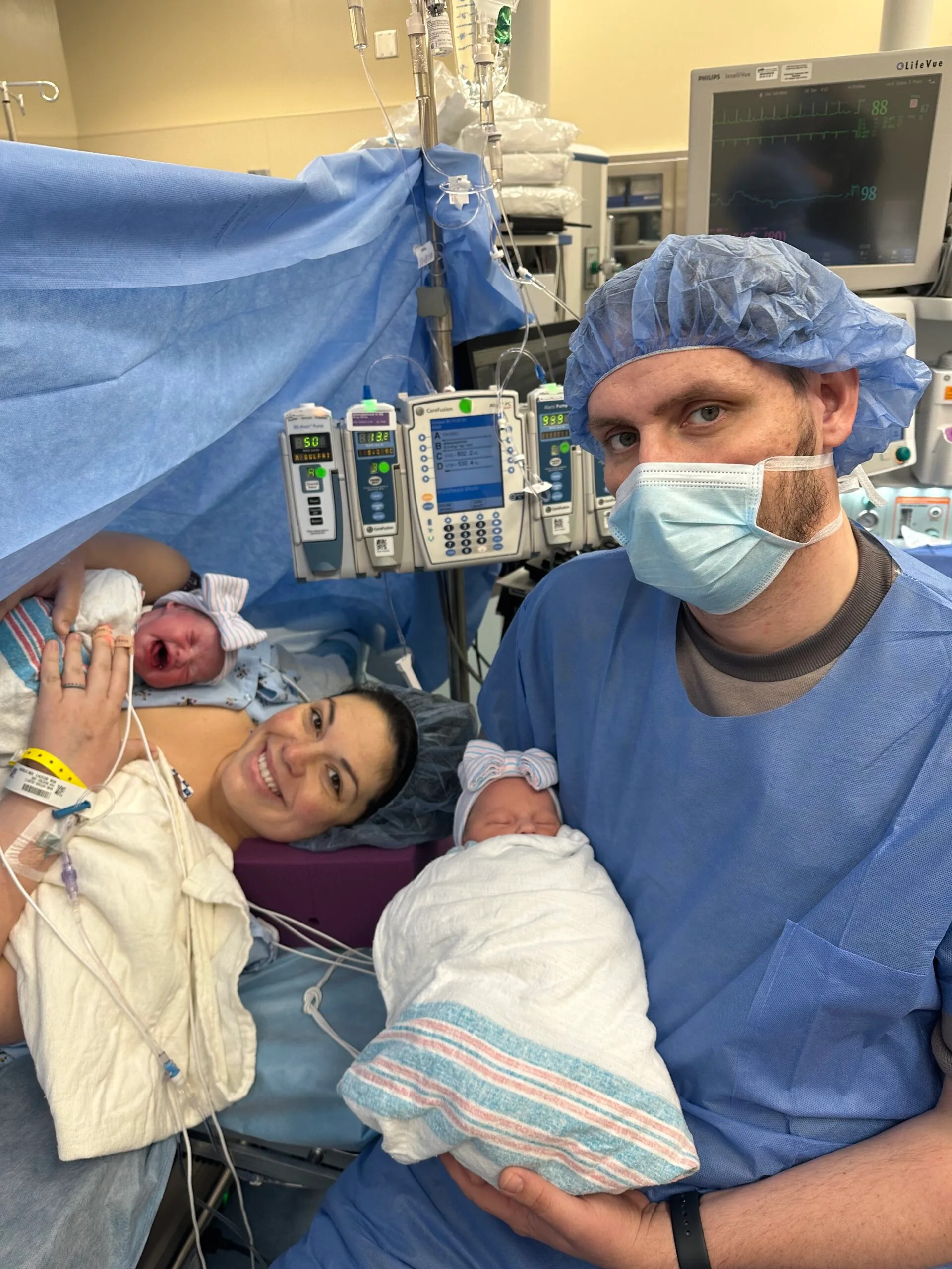 mum and dad in delivery ward wearing scrubs holding two babies  
