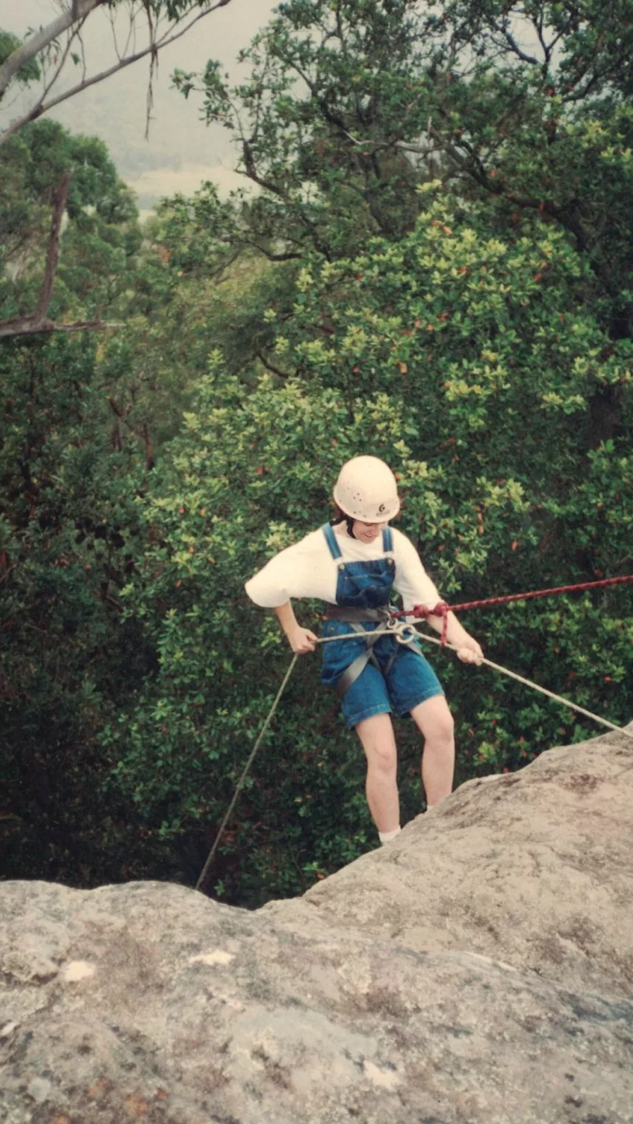 Woman wearing denim overalls and white helmet, strapped up while abseiling.