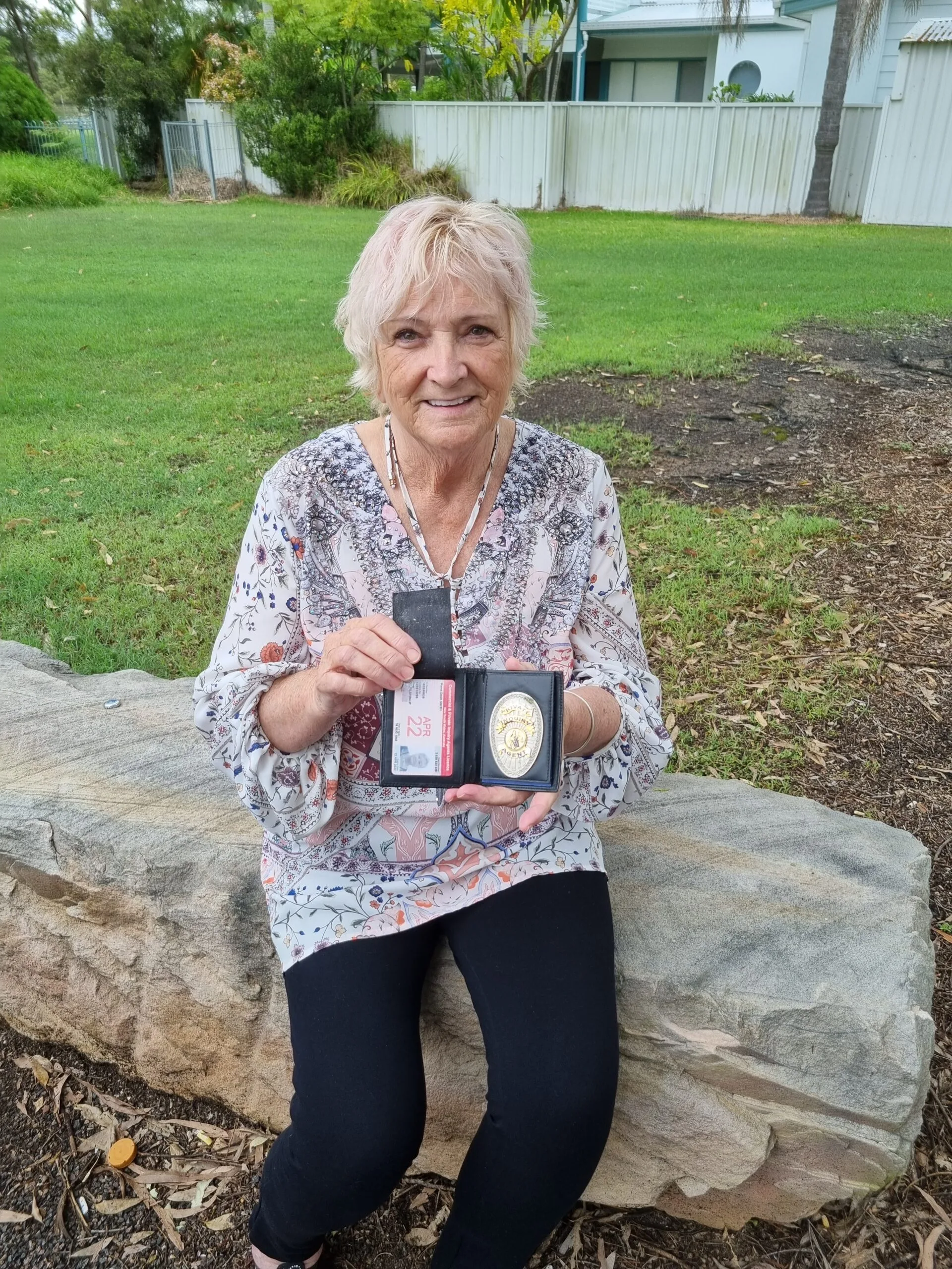 Elderly woman with white hair, wearing a white blouse with floral patters, sitting on a pavement holding up a license and badge.