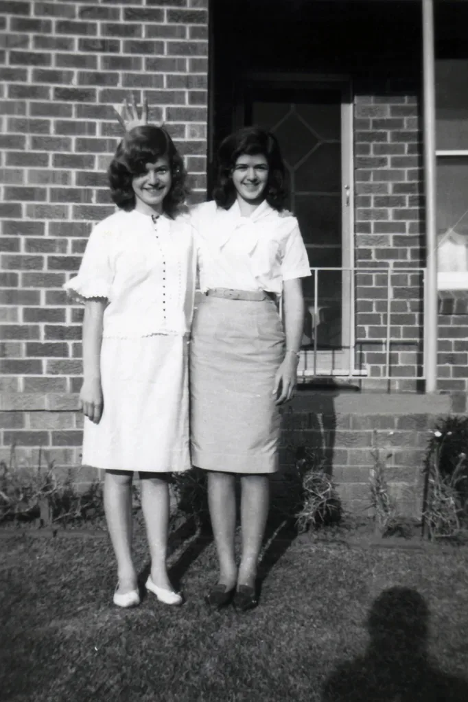 Left: Young girl with short hair wearing white blouse and mid-length skirt wrapping arm around another girl next to her.
Right: Young girl with short brown hair, wearing white blouse and mid length brown skirt smiling at camera.
Black and white photo.