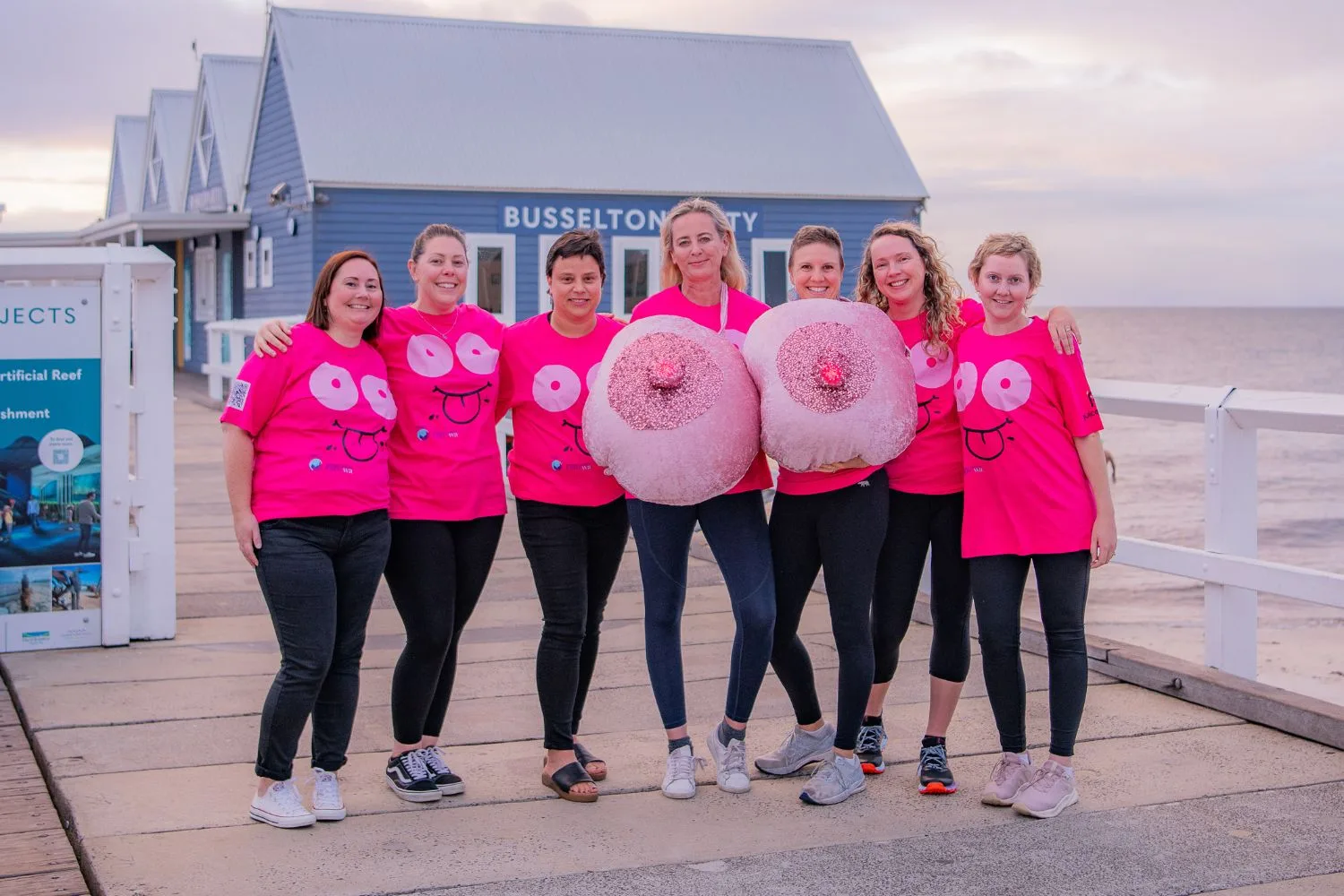 Women standing together in pink tops with inflatable boobs on a boardwalk by water