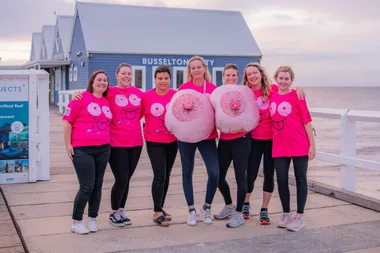 Women standing together in pink tops with inflatable boobs on a boardwalk by water