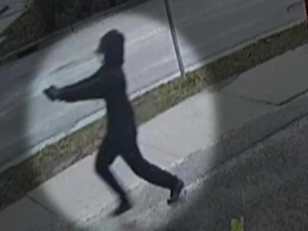 A woman's figure dressed in all black holding up a gun on the footpath.