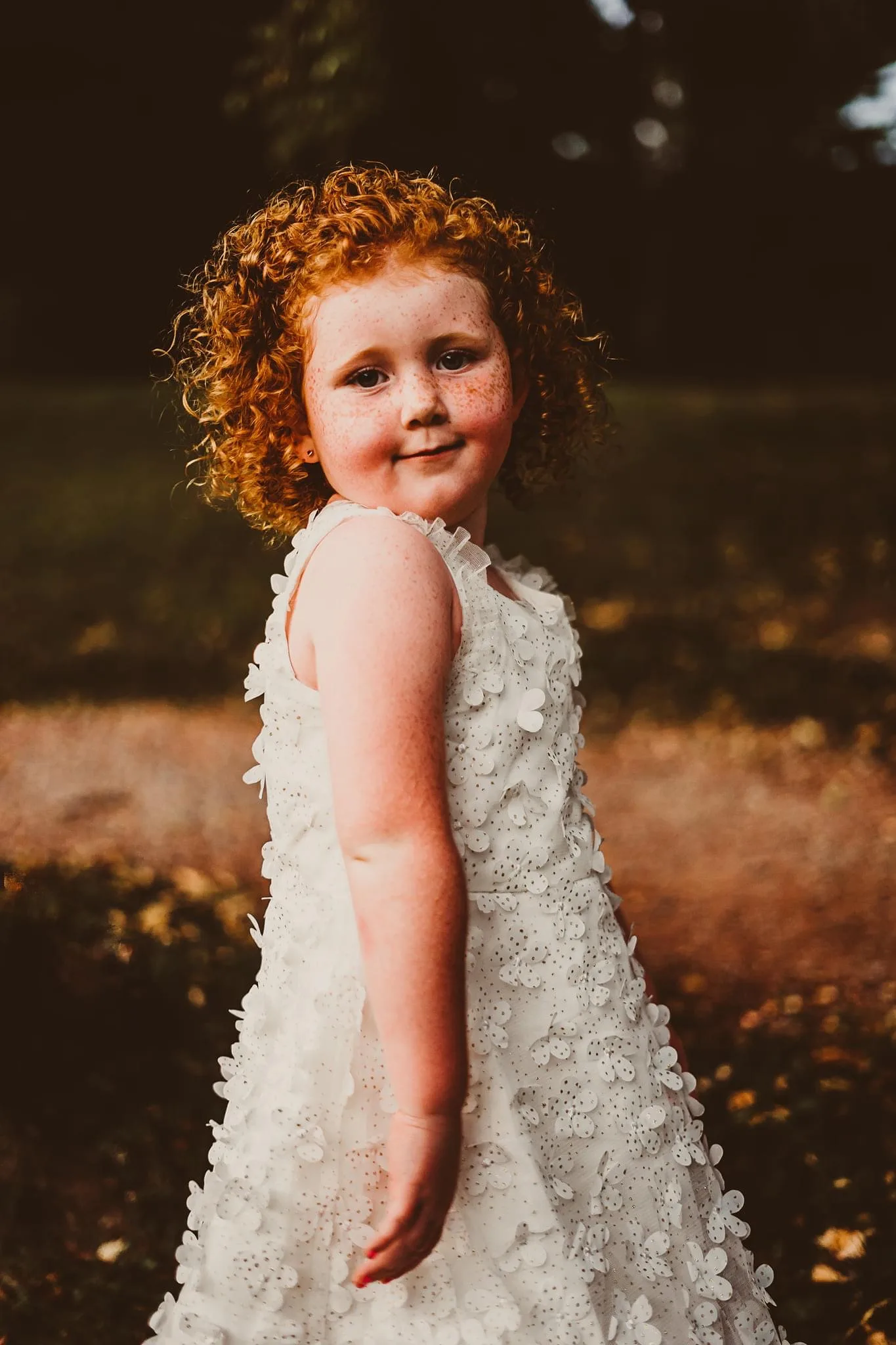 Little girl with bright red hair and freckles across cheeks wearing white dress and smiling at camera.