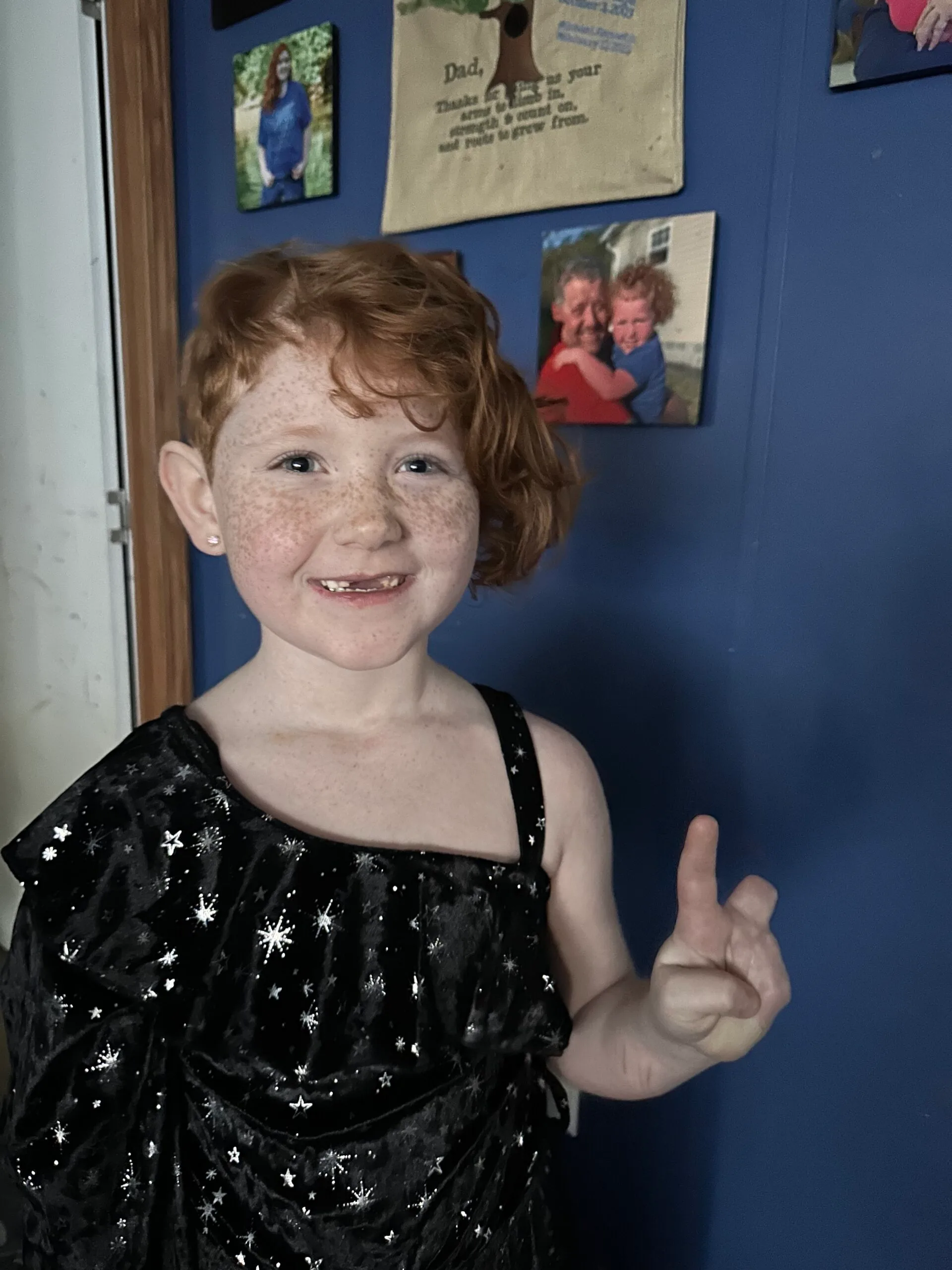 Little girl wearing black dress infront of blue door holding up index finger while thumb bent under. Smiling at camera.