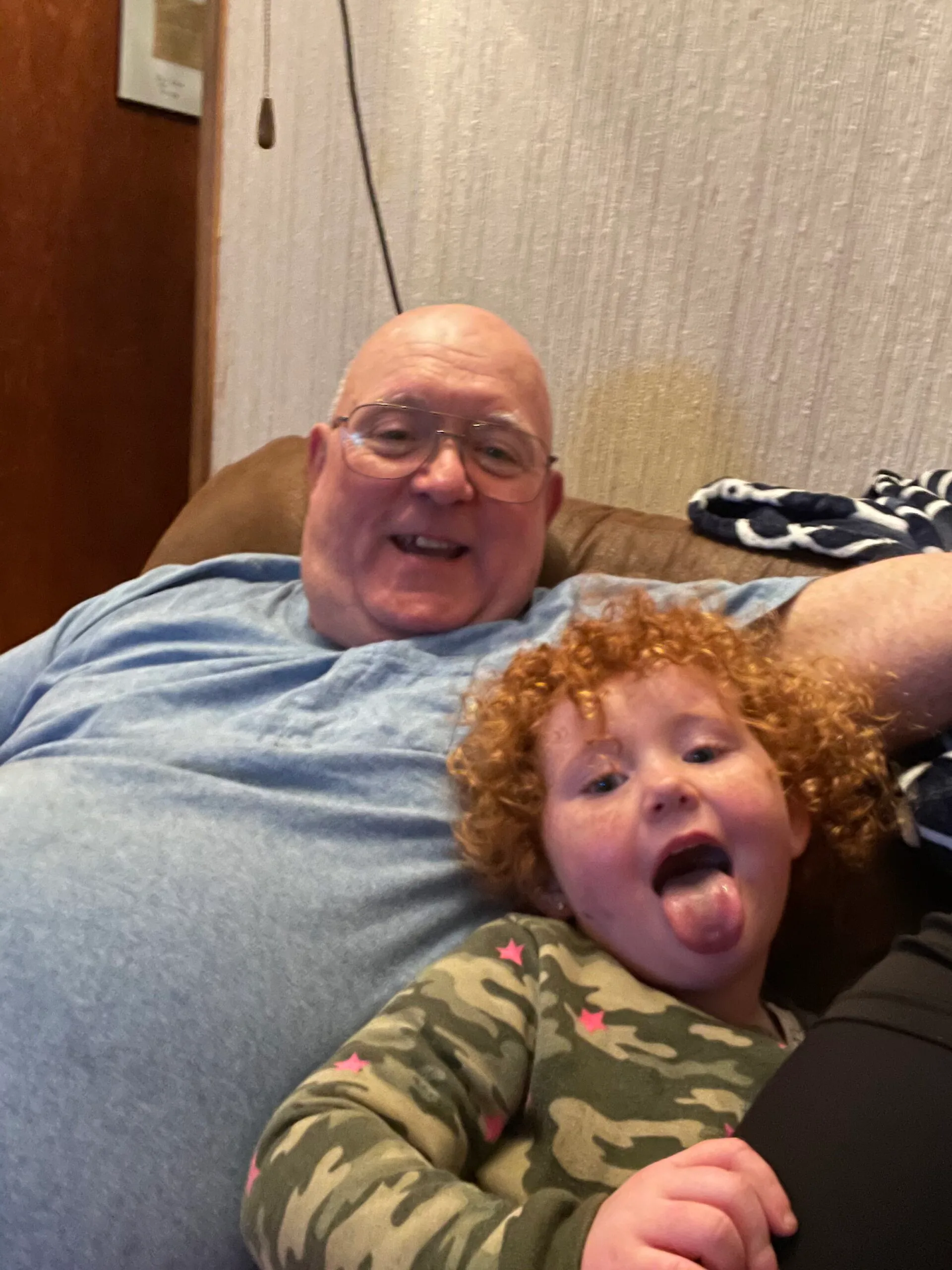 Left: Elderly man wearing glasses and blue shirt, smiling at camera.
Right: Little girl with red hair poking tongue out at camera in elderly mans arm.