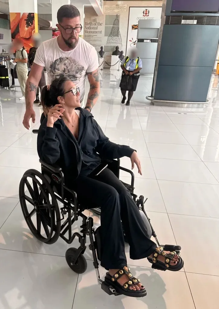 Woman dressed in black tracksuit with brown hair and glasses sitting on wheelchair while man dressed in white shirt behind.
