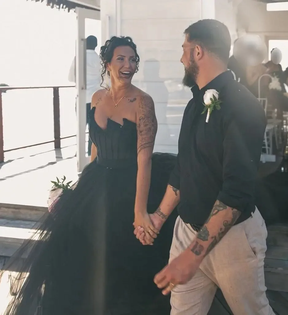 Woman in black wedding dress holding hands with man dressed in black shirt smiling at camera.