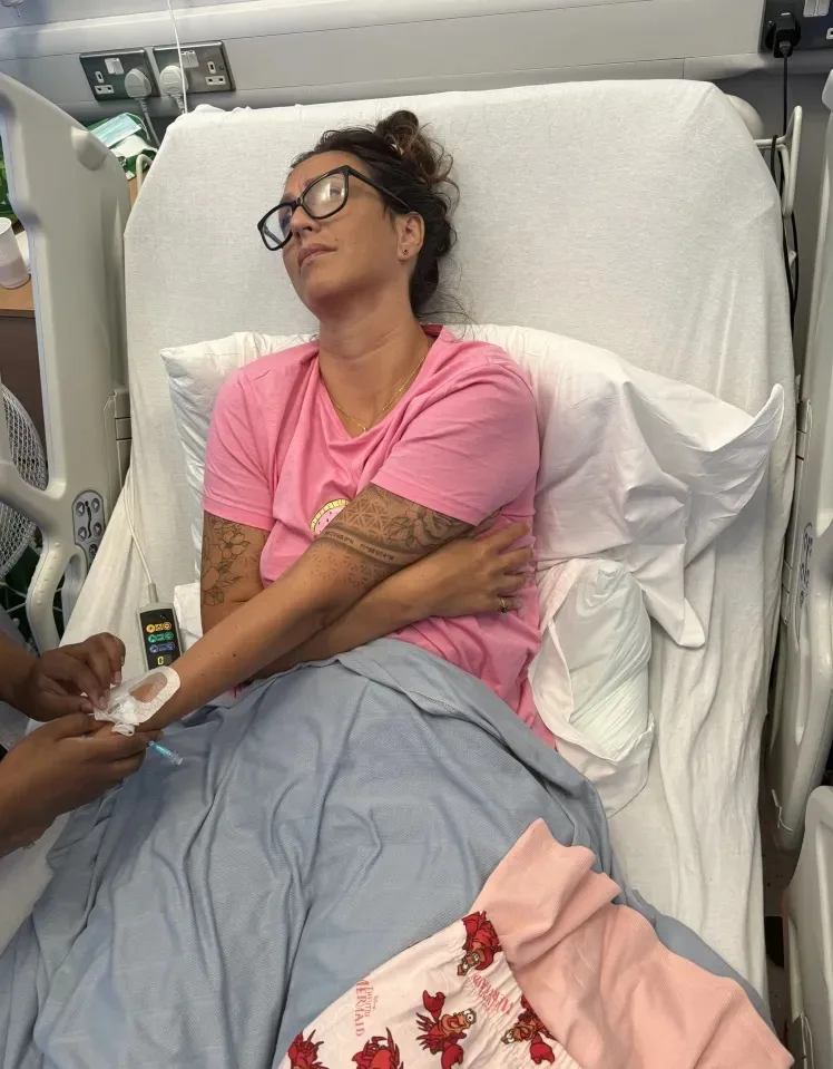 Woman with brown hair wearing glasses and pink shirt looking distressed on hospital bed.