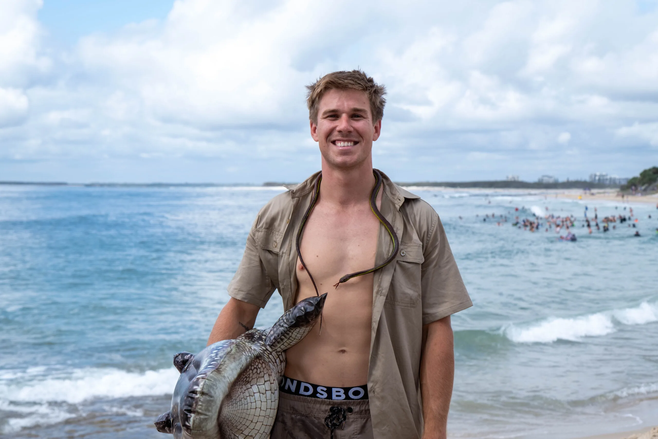Man in khaki shirt with plastic snake around neck holding inflatible croc