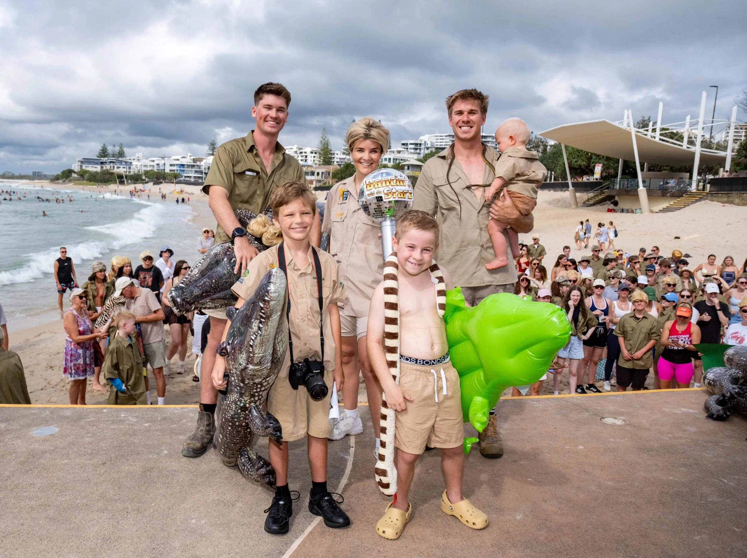 Men, women and children dressed in khaki holding inflatable crocodiles in front of a crowd at the beach