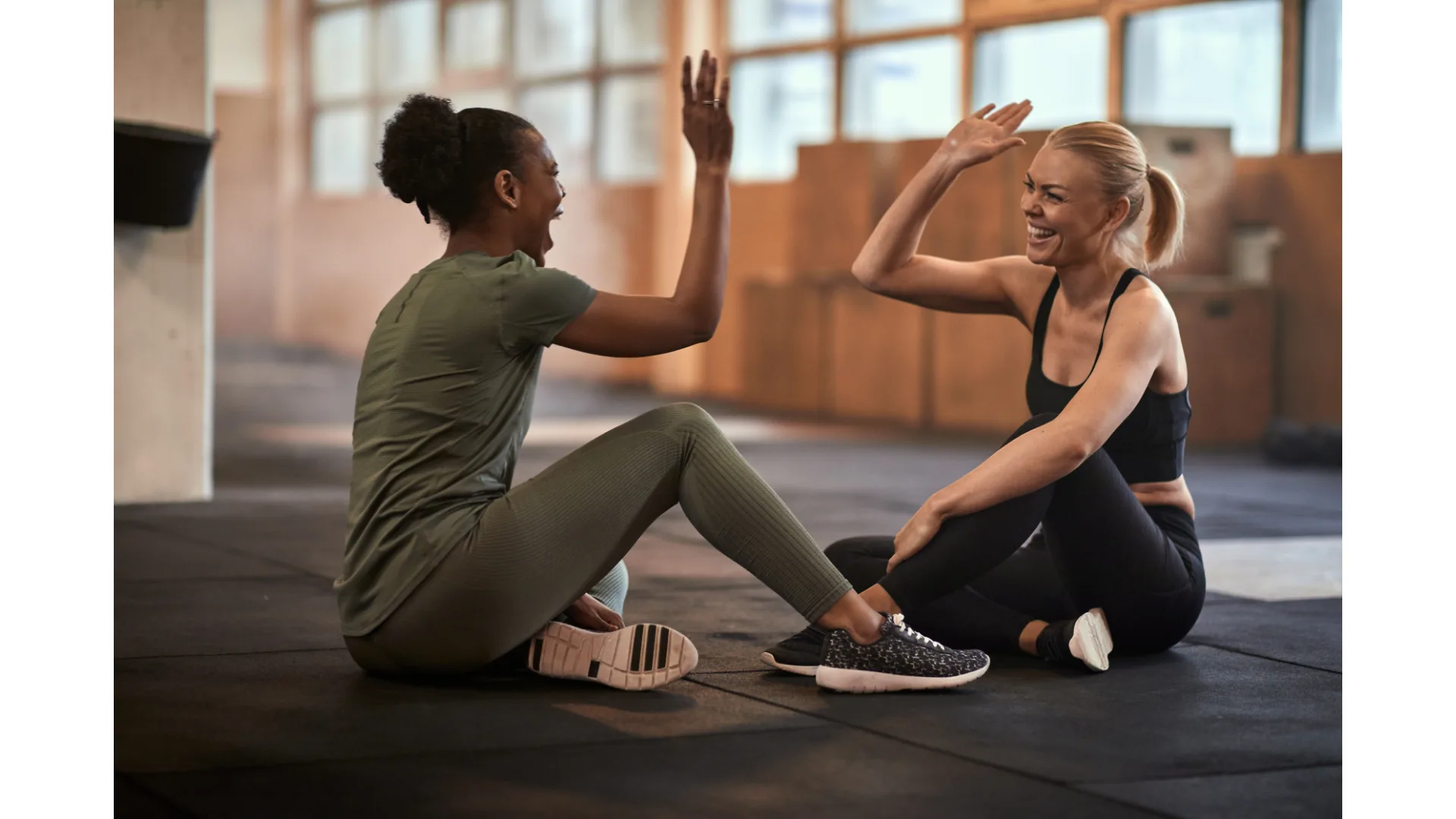 Two friends high five each other while seated on the floor of a gym.