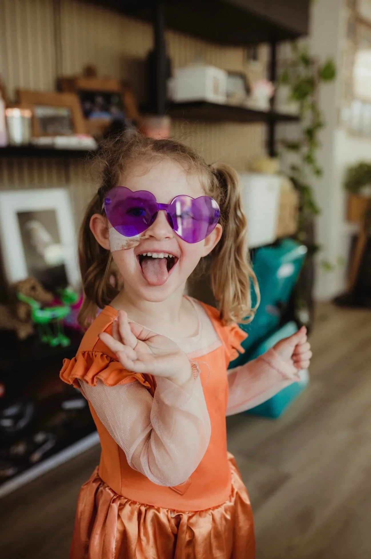 Charlotte wearing an orange dress and purple coloured heart-shaped glasses. She is smiling with her tongue out