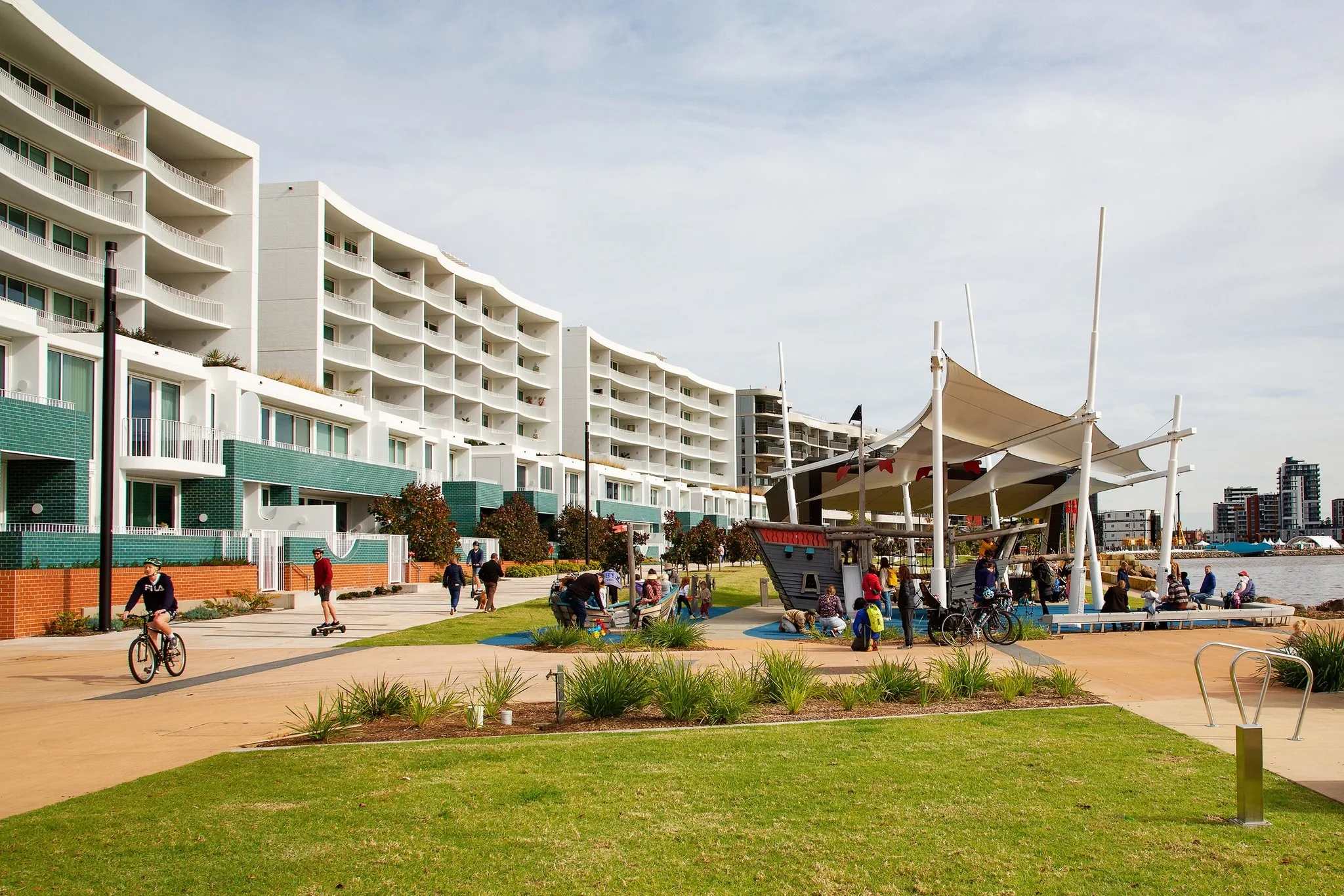 Picture of park, children riding on bikes, grass and playground structures near waterfront.