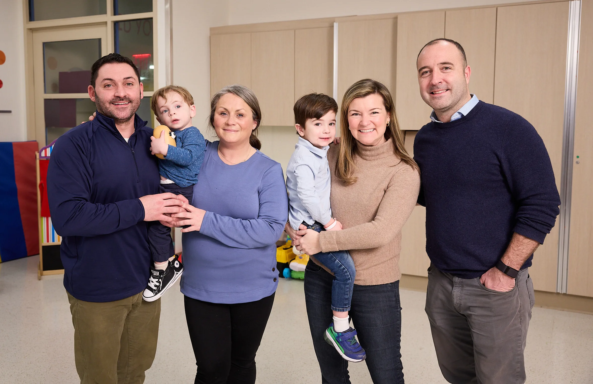 Left: Man dressed in blue shirt holding baby alongside women dress in blue top.
Right. Woman in brown turtle neck top holding toddler in arms next to man dressed in dark blue sweater,.