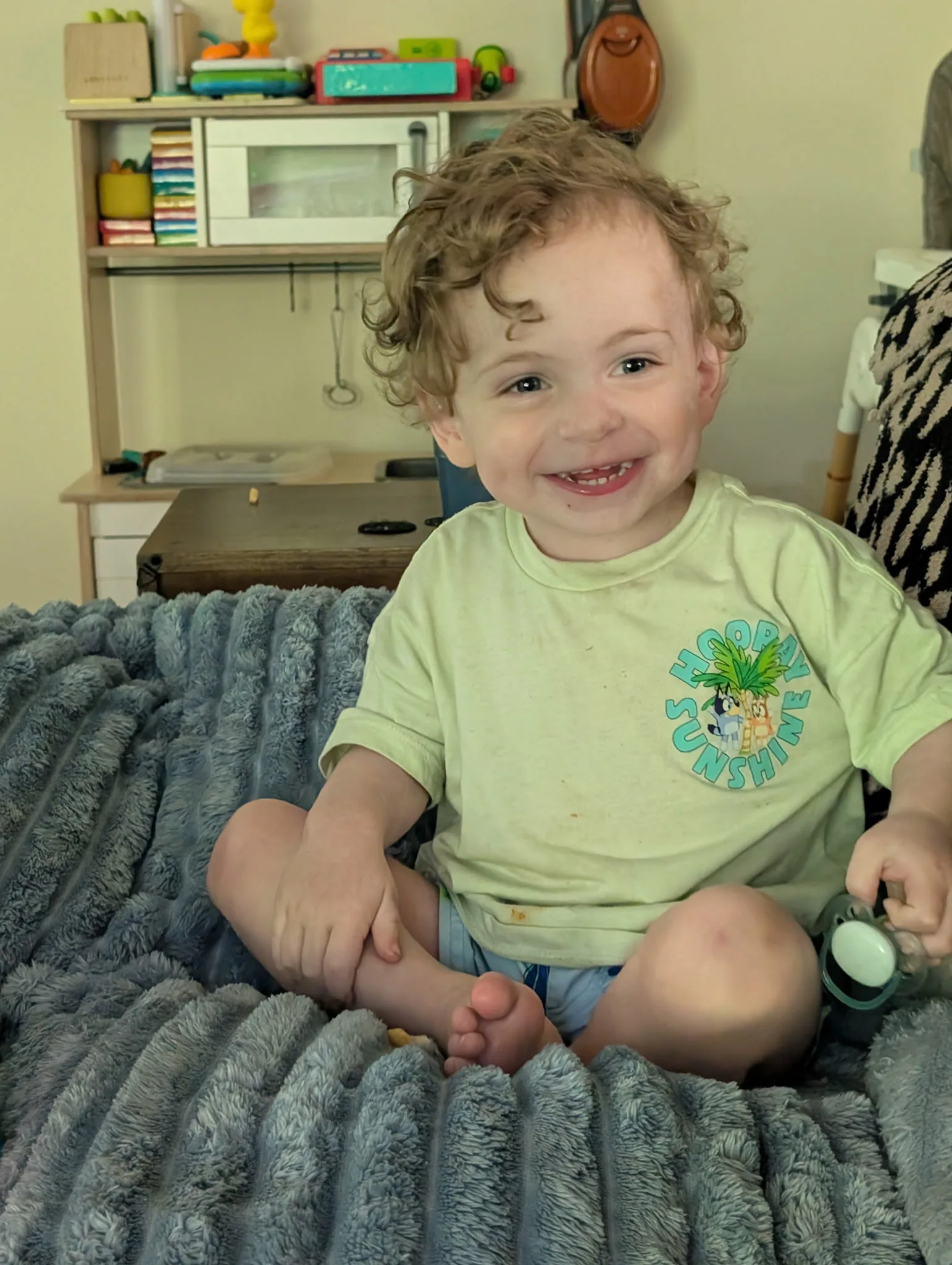Toddler in yellow shirt and blue shorts sitting on top of bed smiling at camera.