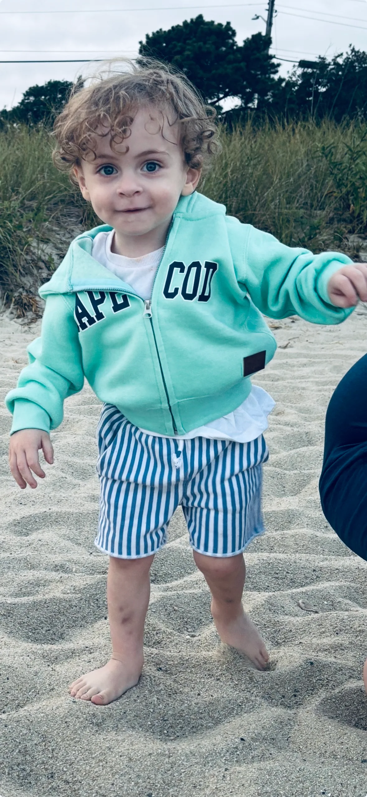 Toddler with blonde curls, wearing blue jacket standing on sand and smiling at camera.
