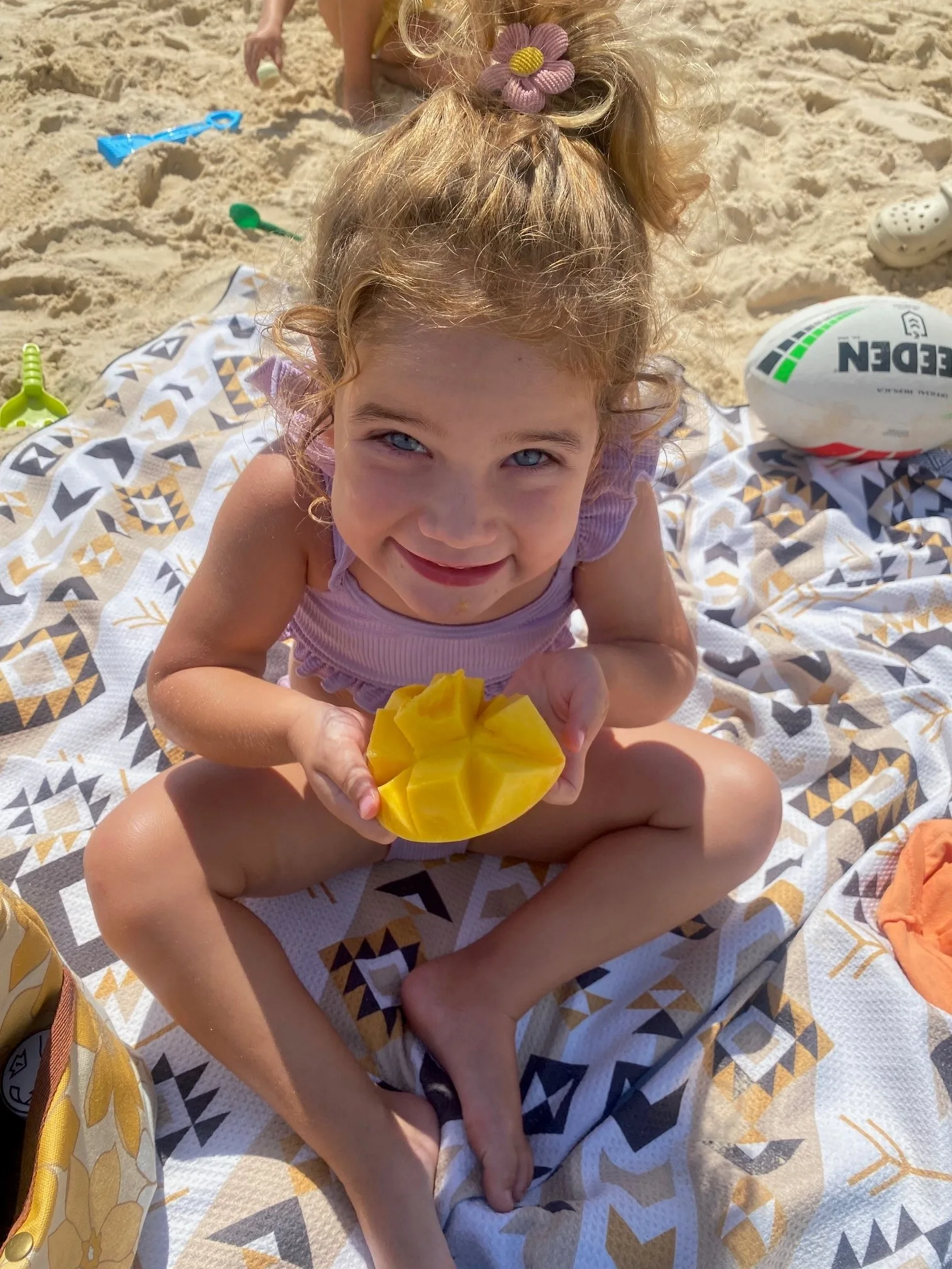 Little girl with blonde hair wearing purple top, holding a mango and sitting on sand at the beach. Smiling at camera.