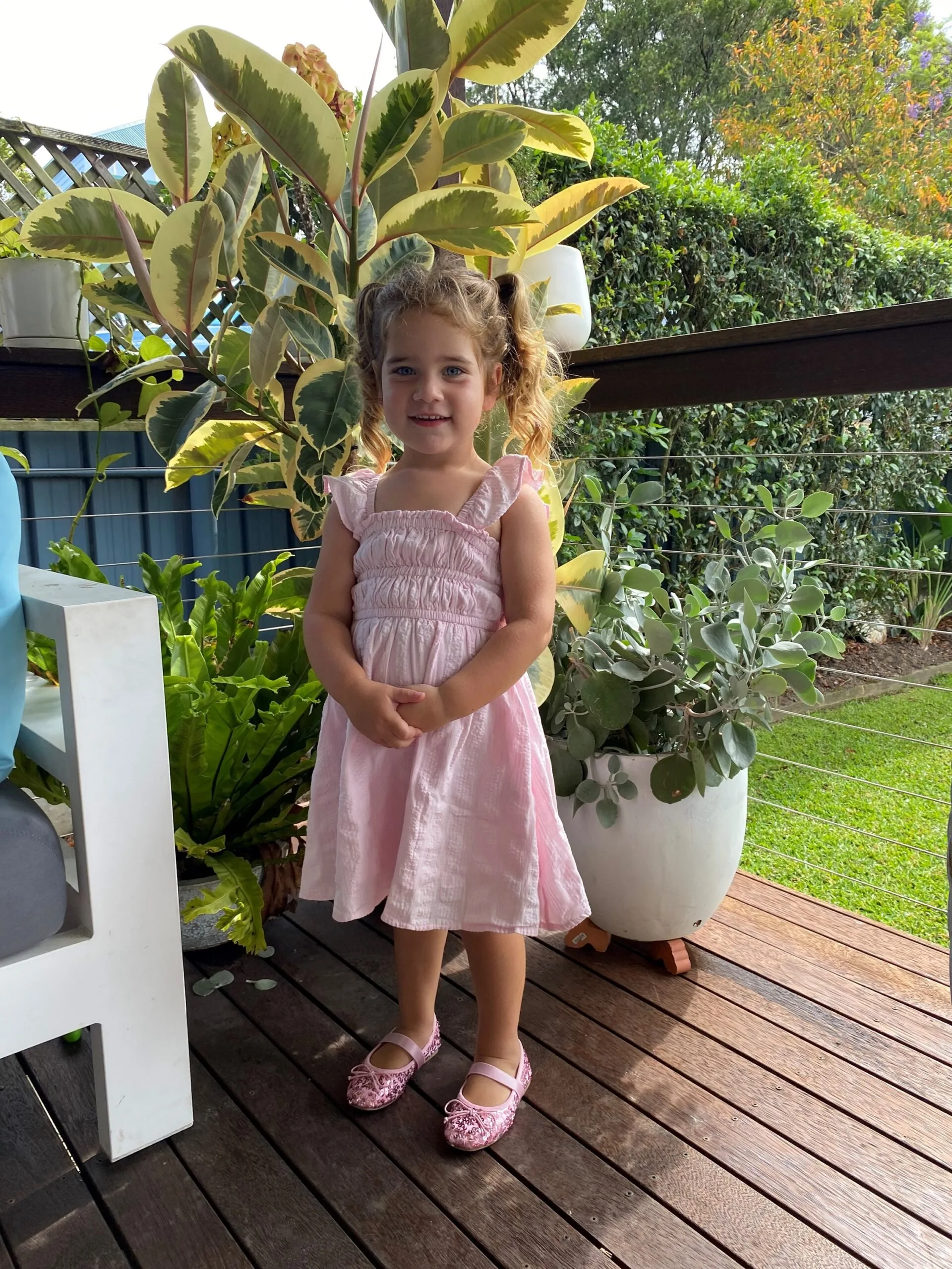 Little girl with two blonde piggy tails, blue eyes, wearing pink dress and standing with arms crossed and smiling at camera.