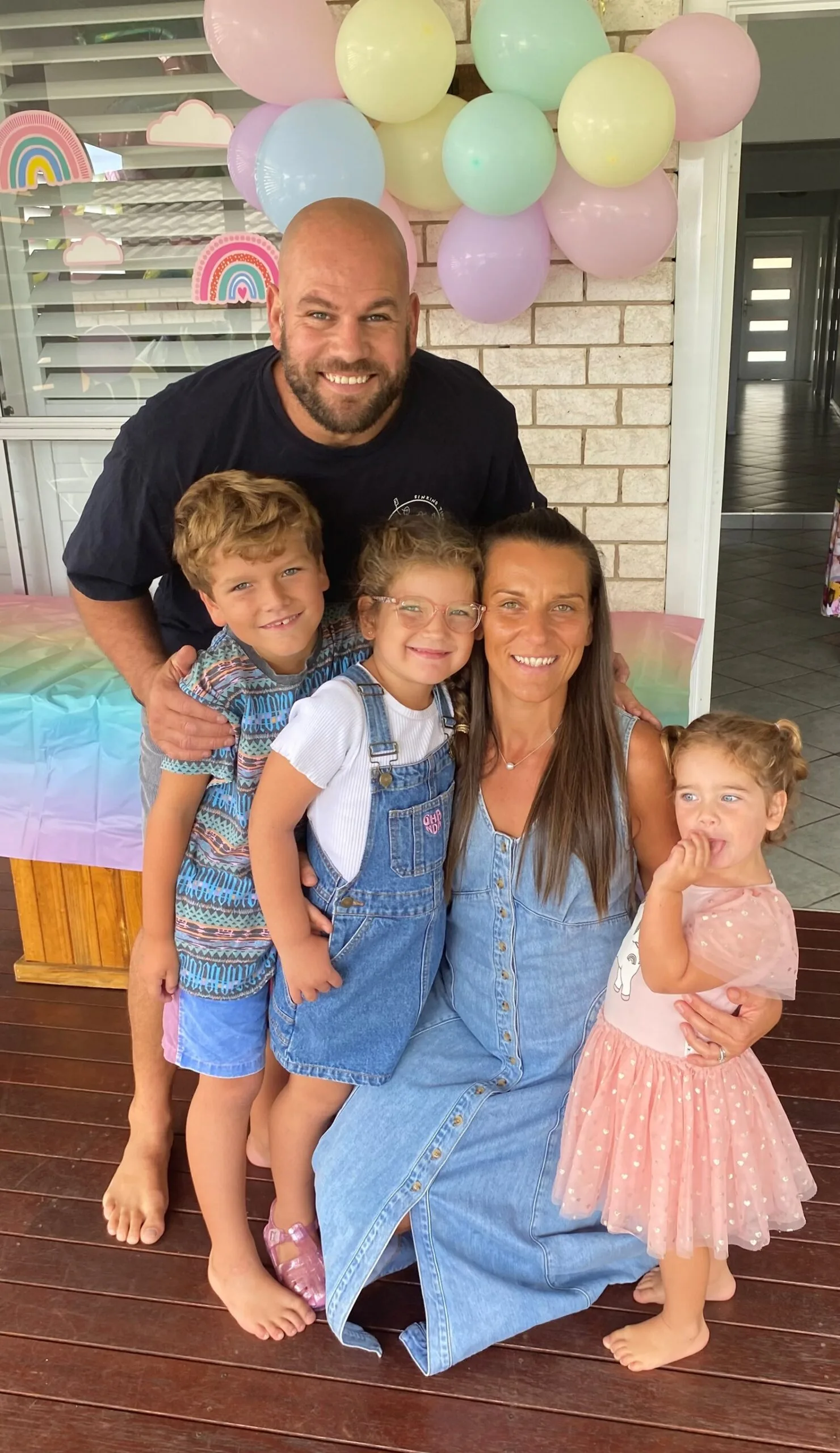 Man wearing black shirt wraps arms around little boy with blonde hair standing next to little girl with blonde hair and glasses who is standing next to woman wearing long blue dress and wrapping arm around baby girl wearing pink tutu dress. All smiling at camera.