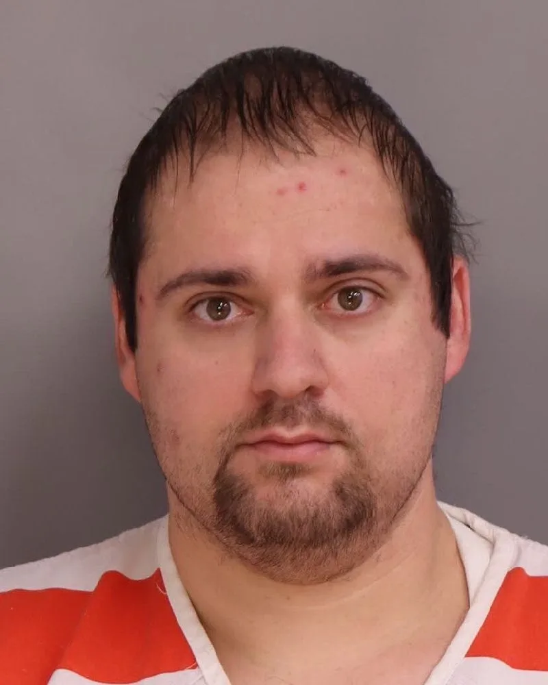 Man with brown hair and beard staring into camera wearing orange and white striped shirt.