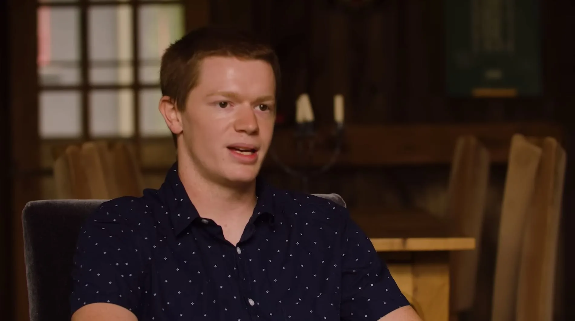 Man with brown hair sitting on chair wearing blue shirt with white dots speaking to camera.