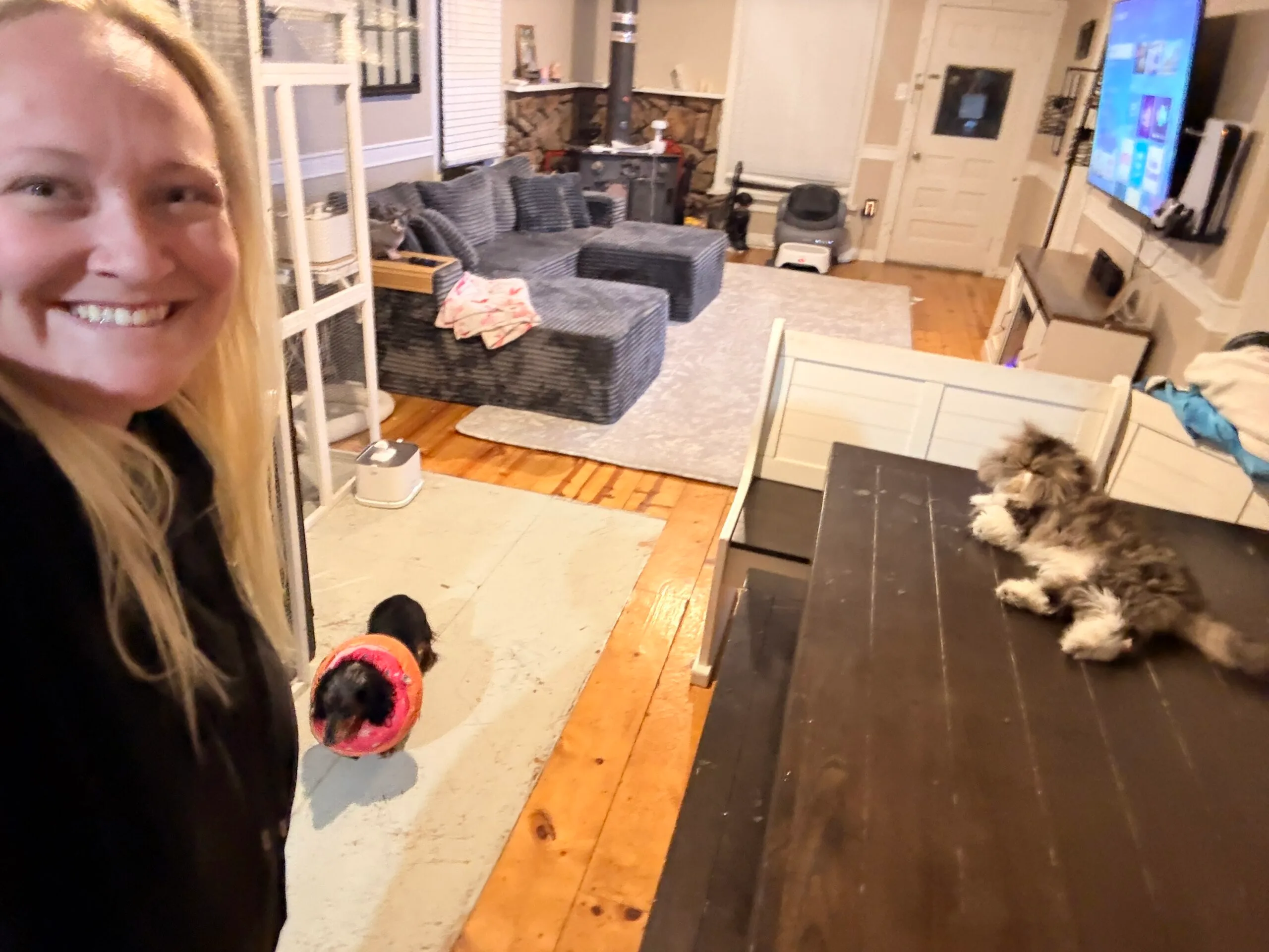 Woman with blonde hair wearing black shit smiling next to clean living room and dinning table.