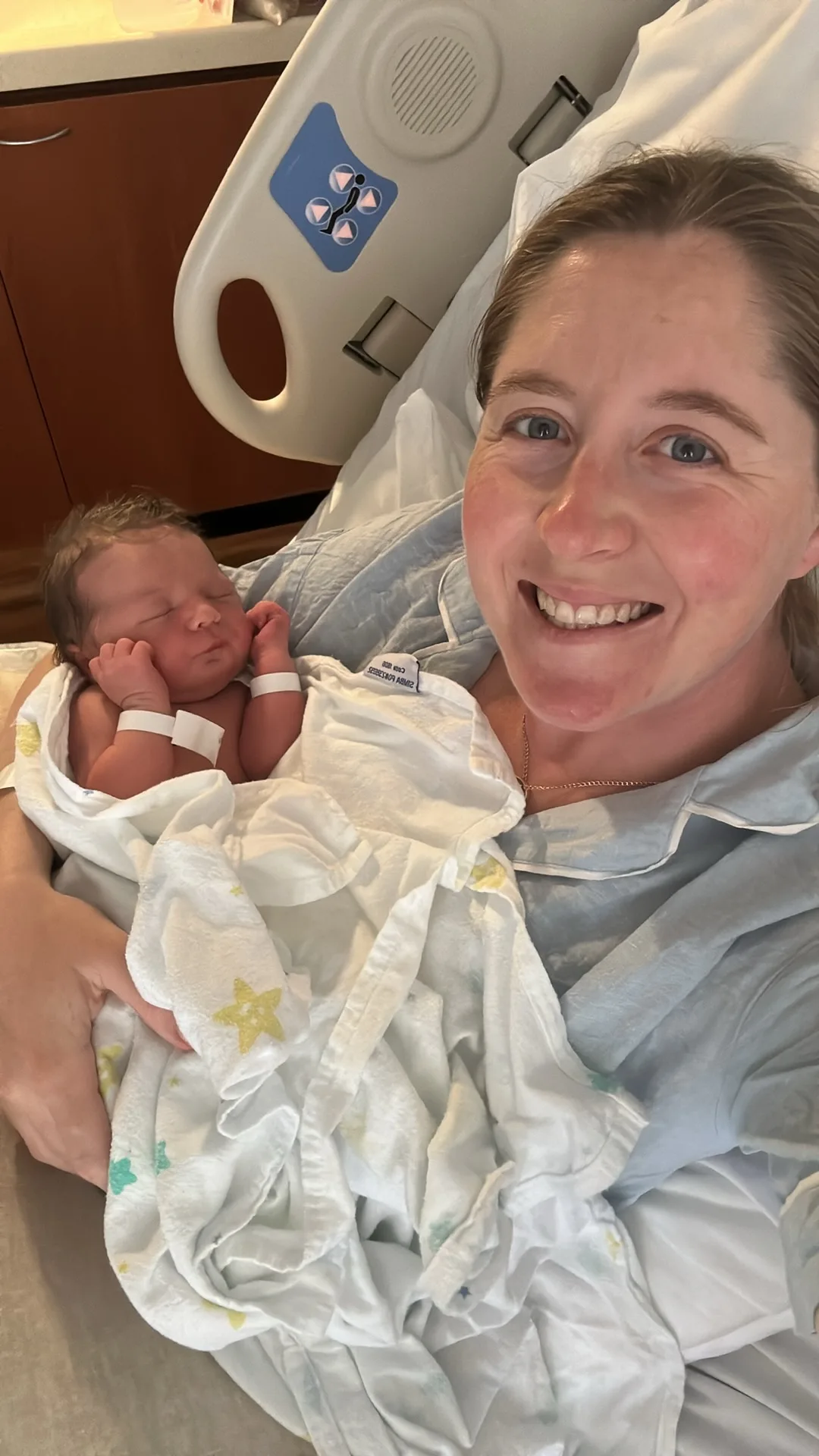 Woman on hospital bed holding newborn baby and smiling at camera.