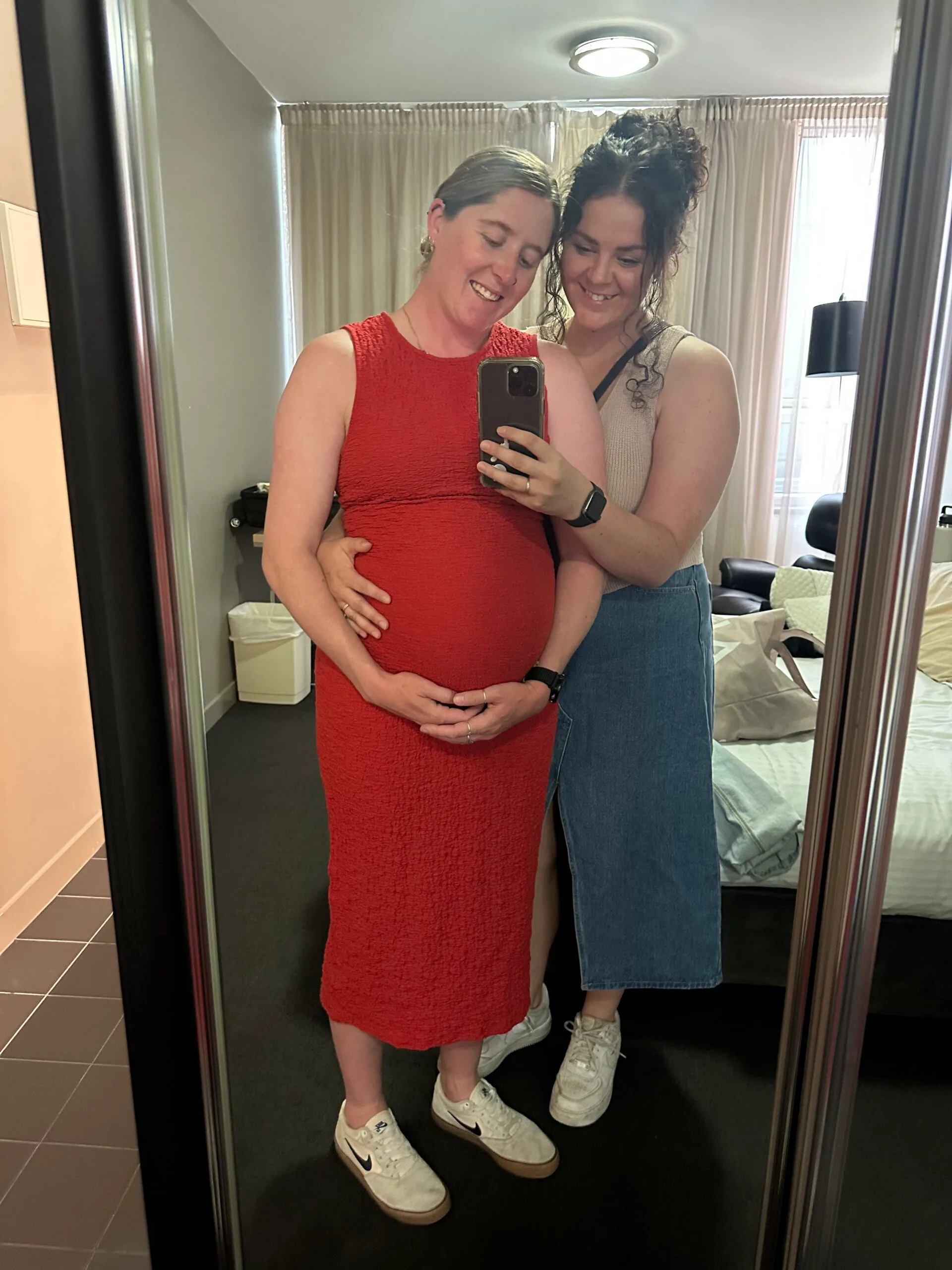 Left: Pregnant woman with blonde hair tied back in red dress holding up bump in the mirror.

Right: Woman with dark curly hair wearing brown shirt wrapping arms around pregnant woman.

Both smiling in the mirror.
