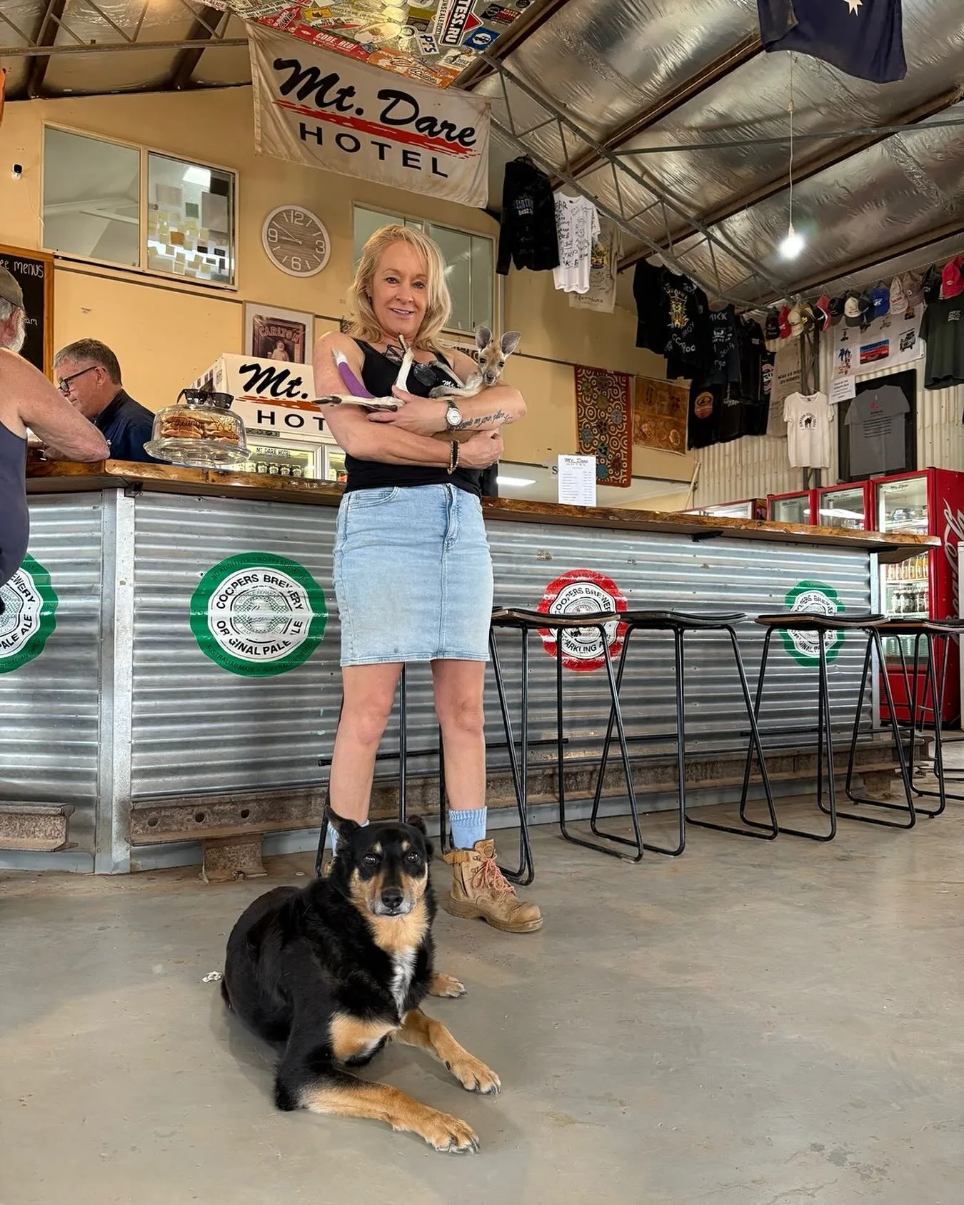 A woman holding a joey kangaroo with a kelpie at her feet, standing inside an outback Australian pub