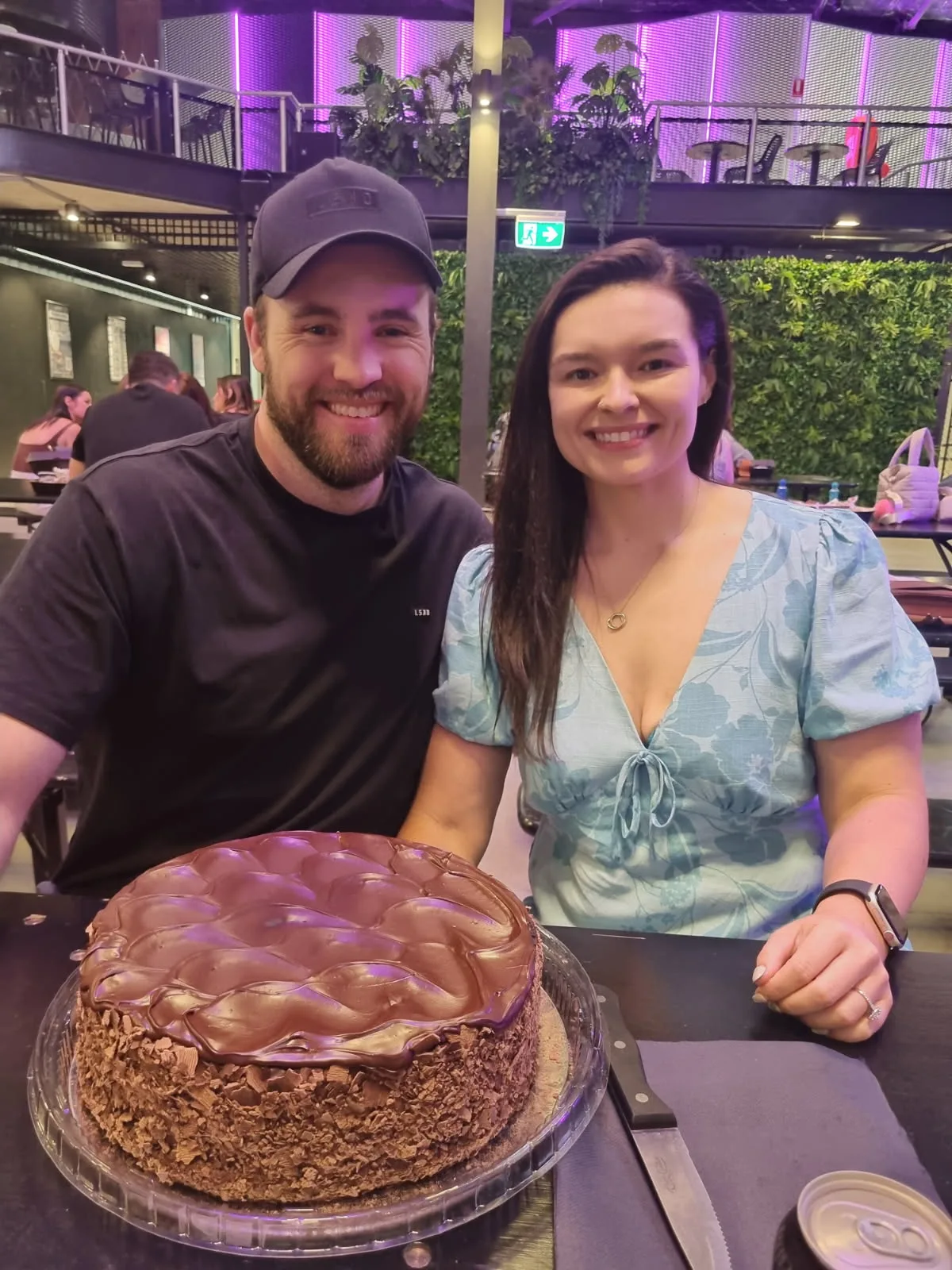woman in blue dress and man in black shirt smiilng in front of cake 