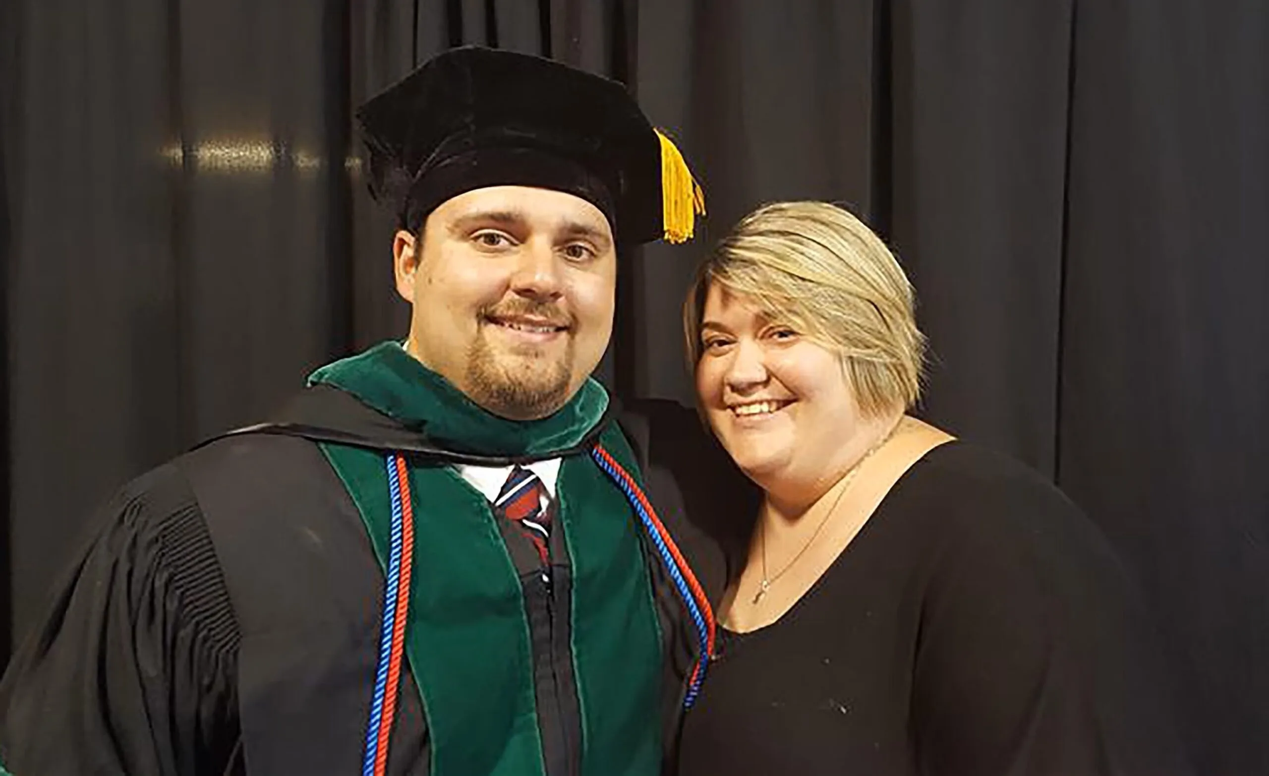 Man in graduating cape and hat smiling at camera while standing next to blonde woman in black top.