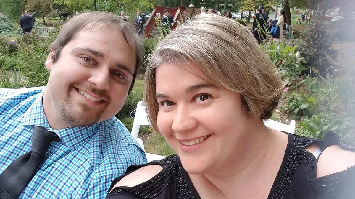 Man with brown hair wearing blue stripped shirt smiling next to woman with short blonde hair wearing black top.