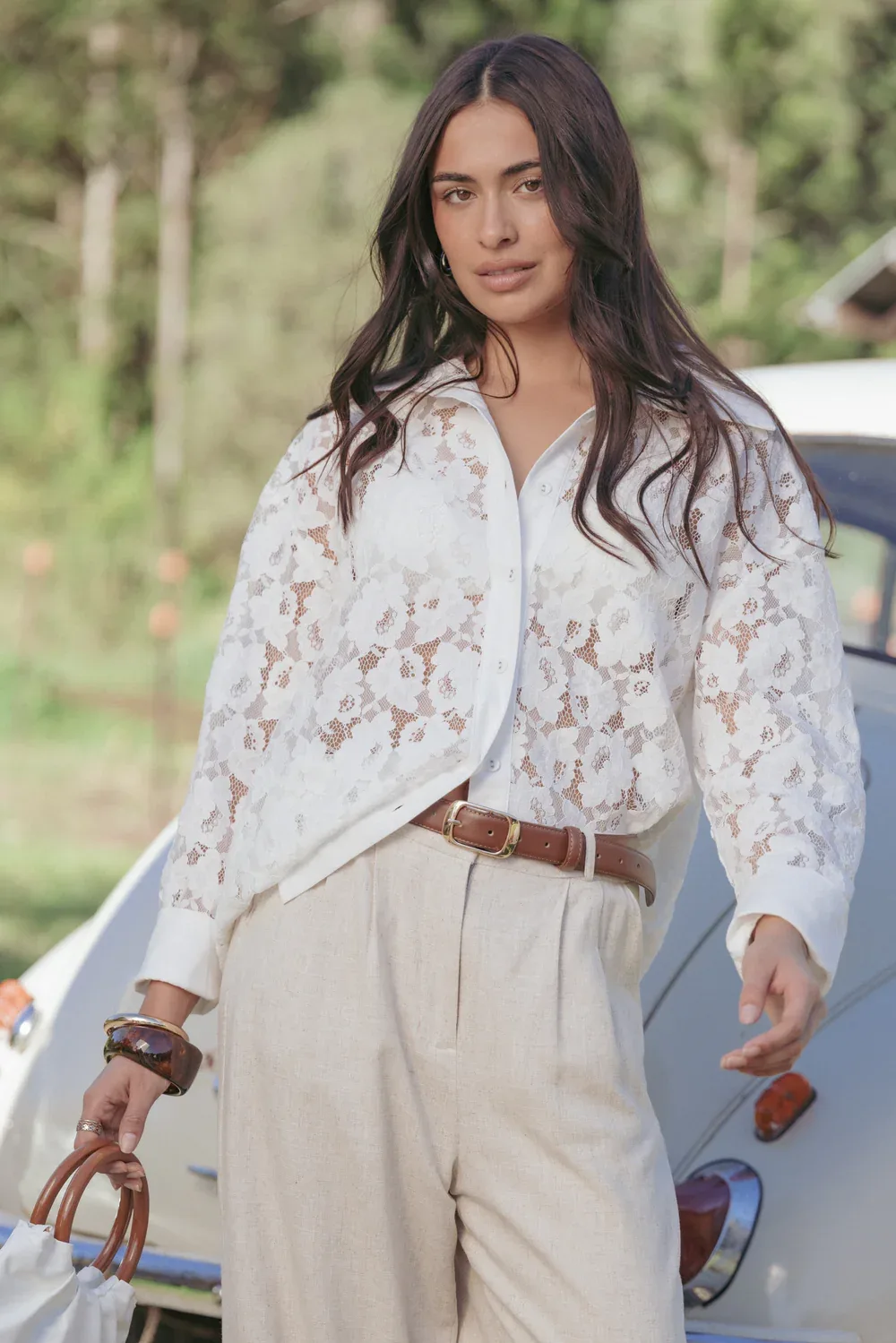 Woman standing near a vintage car, wearing a white lace button-down shirt and relaxed linen trousers