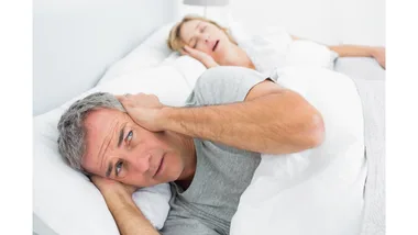 A man covers his ears, while a woman snores next to him in bed.