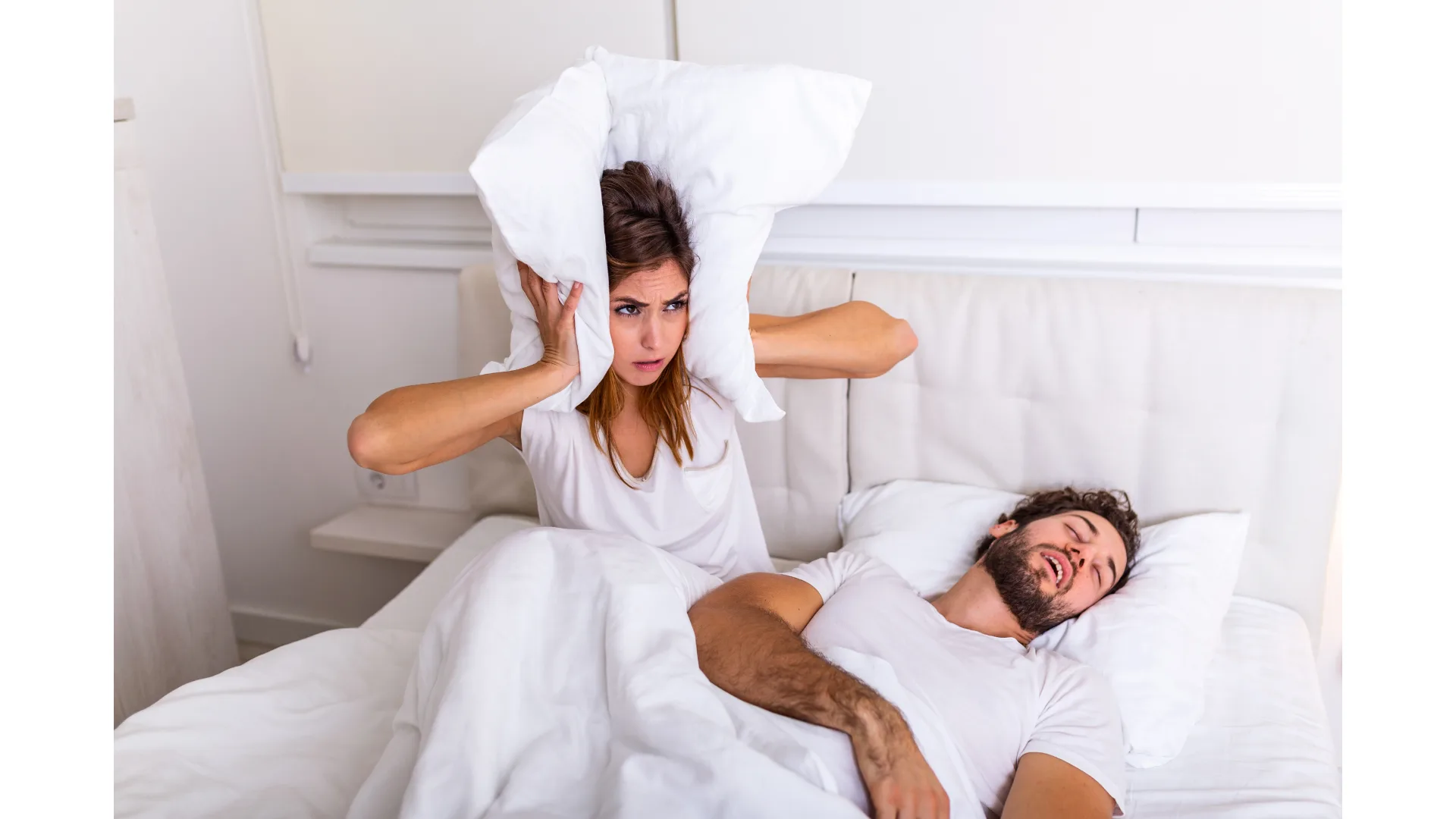 A woman sits up in bed and covers her ears with a pillow while a man sleeps next to her.