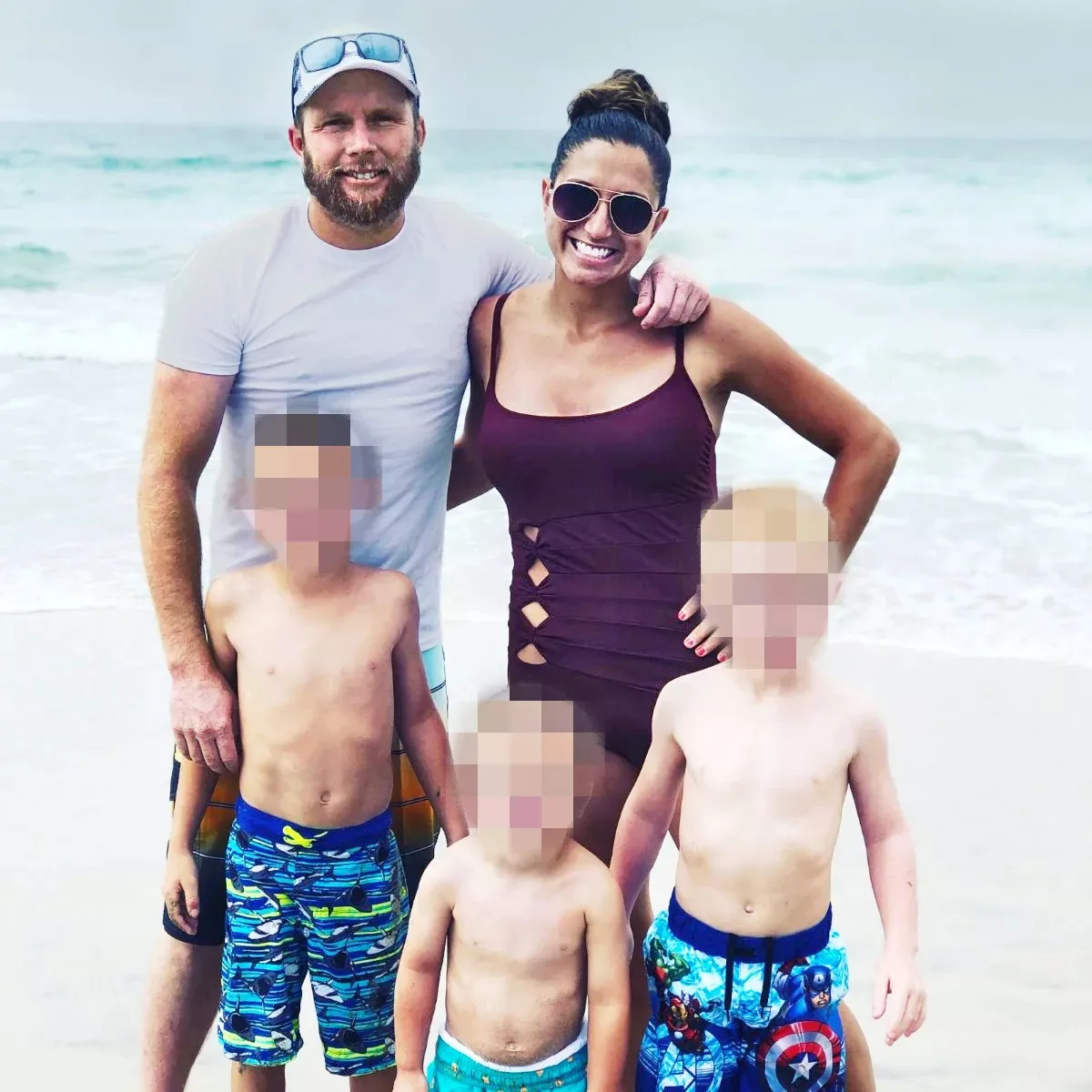 Man wearing great shirt and hat wrapping arms around woman wearing purple bathing suit and sunglasses standing behind three young boys. All are at the beach.