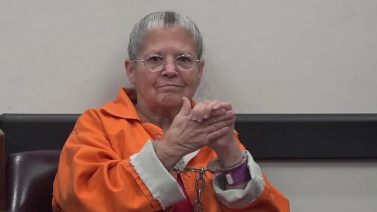 Elderly woman in orange prison suit sitting down with hand cuffs on staring at camera.