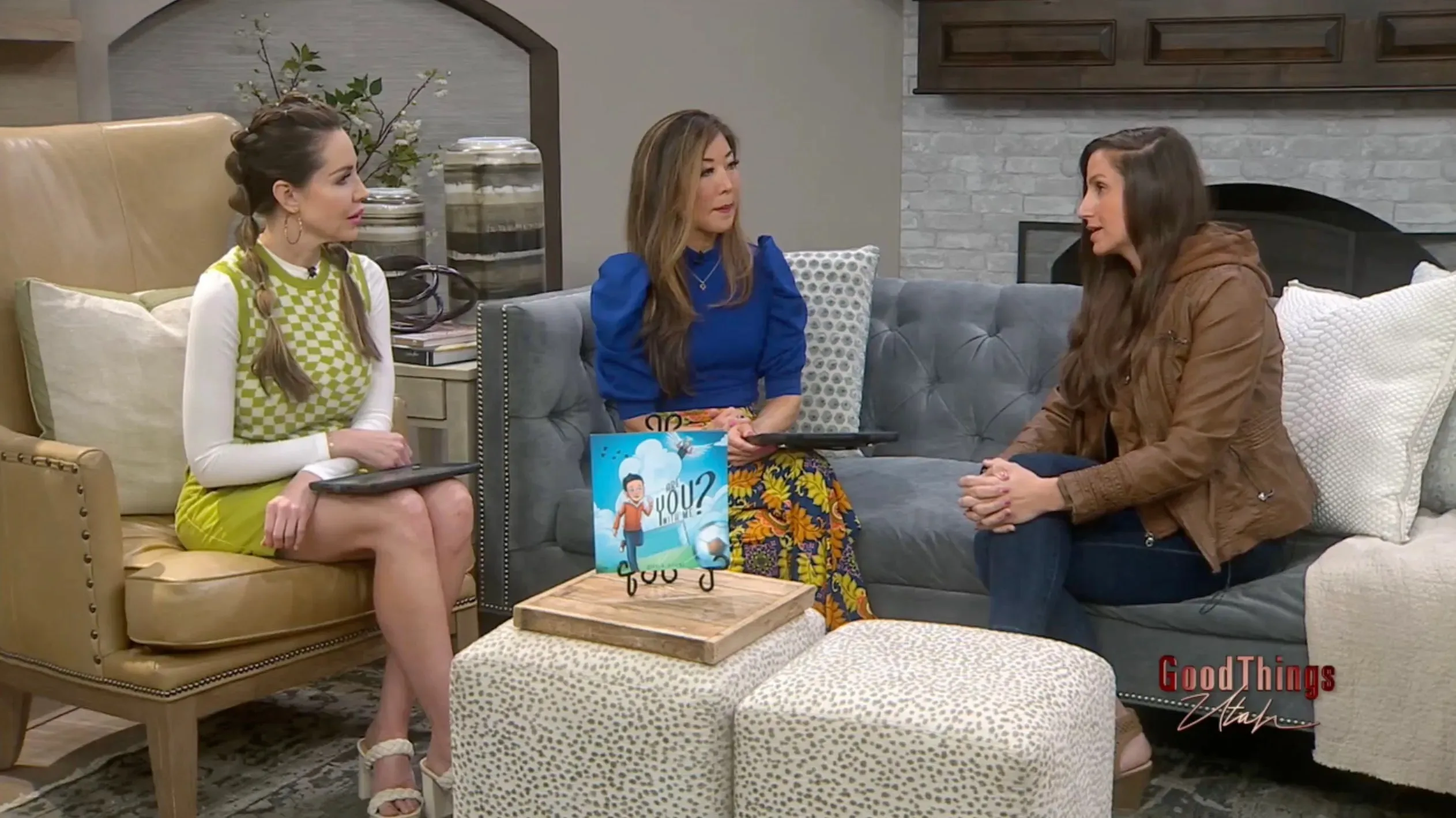 Three women sitting on grey couch facing each other in conversation with blue book on table. 