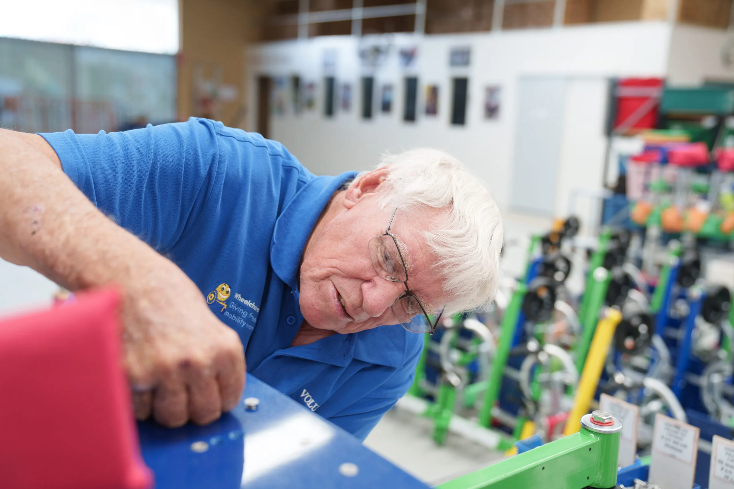 Elderly man in blue collared shirt, wearing glasses and screwing nail on to steel in a workshop. 