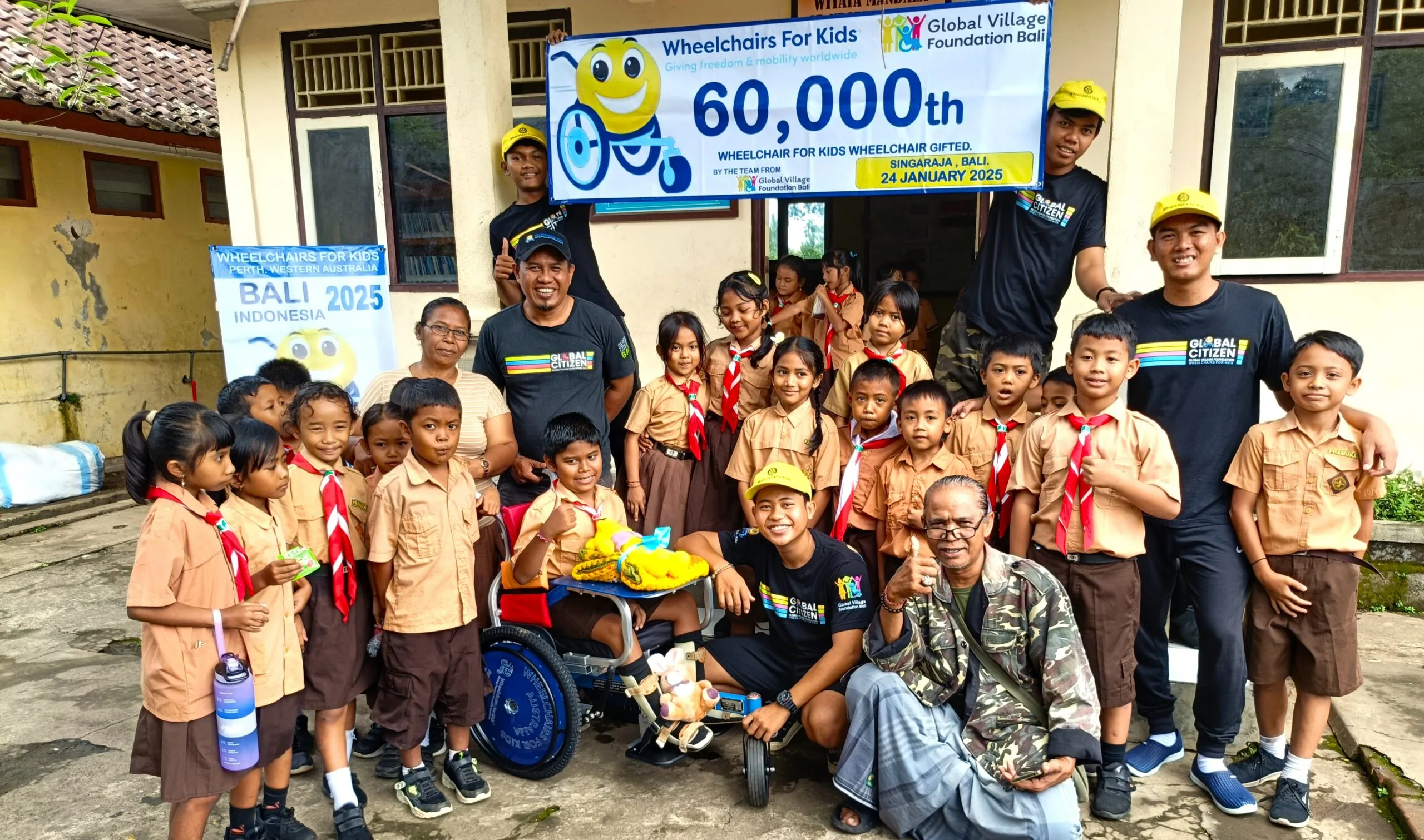 Several kids in brown school uniform smiling at camera while five men wearing black uniforms holding up a sign. One child sitting in a wheelchair.