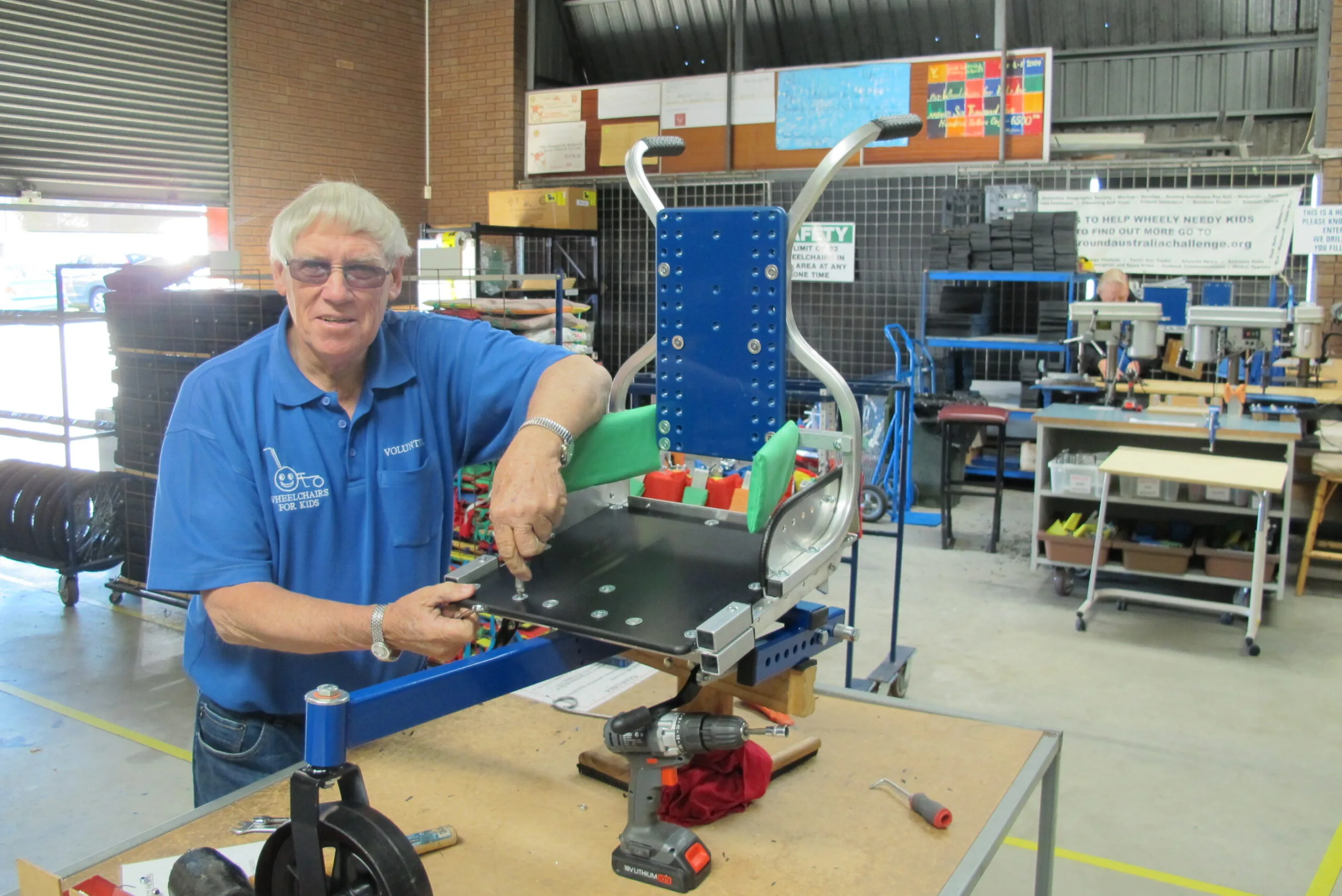 Man wearing blue collared shirt and glasses working on a makeshift wheelchair in a metal workshop. 