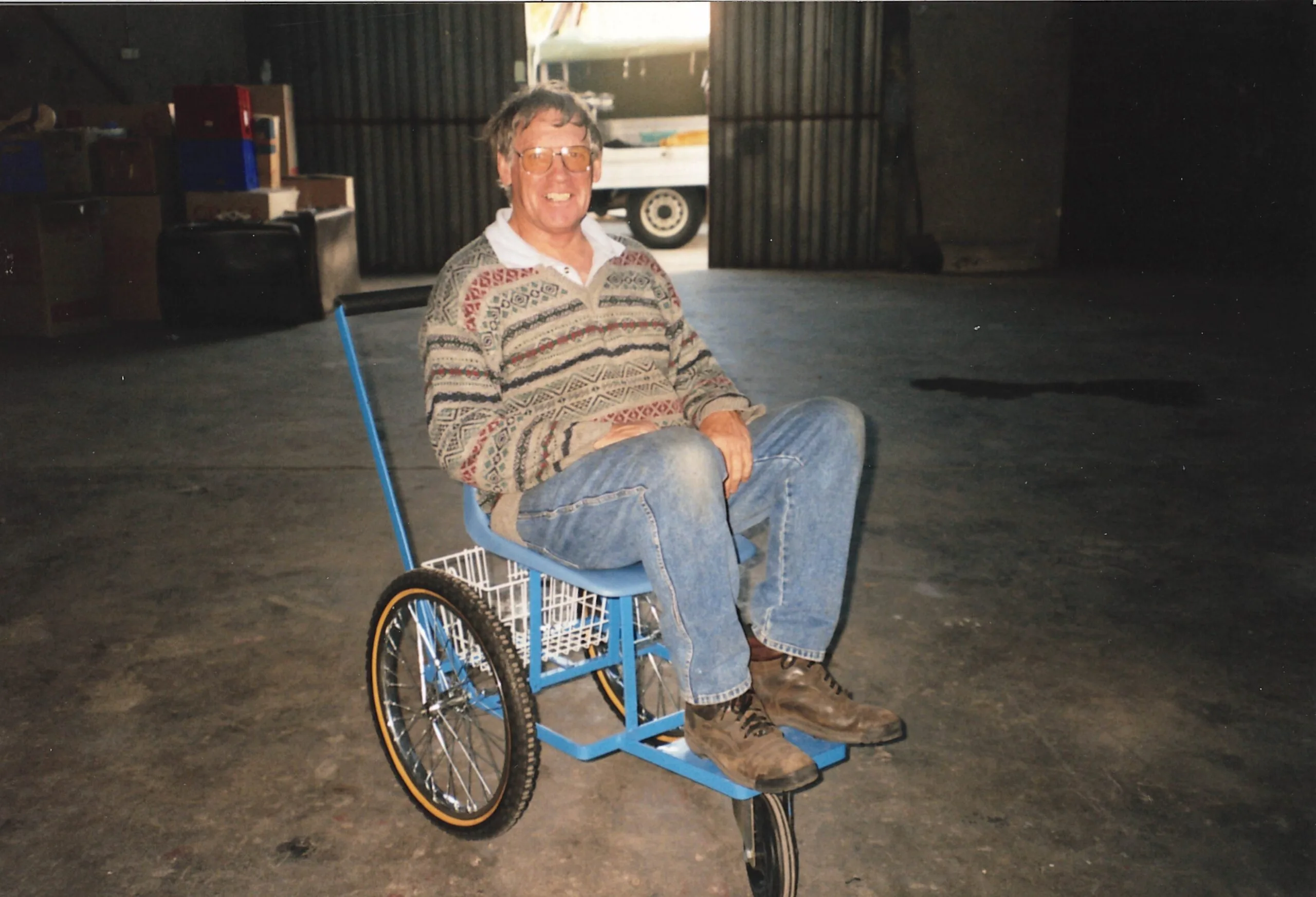 Man wearing brown sweater and jeans sitting on a makeshift wheelchair outside a warehouse.
