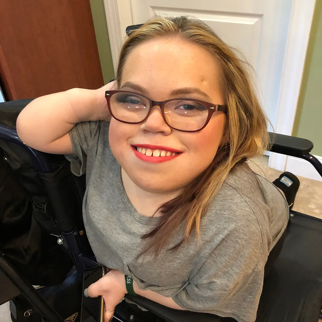 Woman with blonde hair and glasses, wearing grey shirt and smilling at camera.