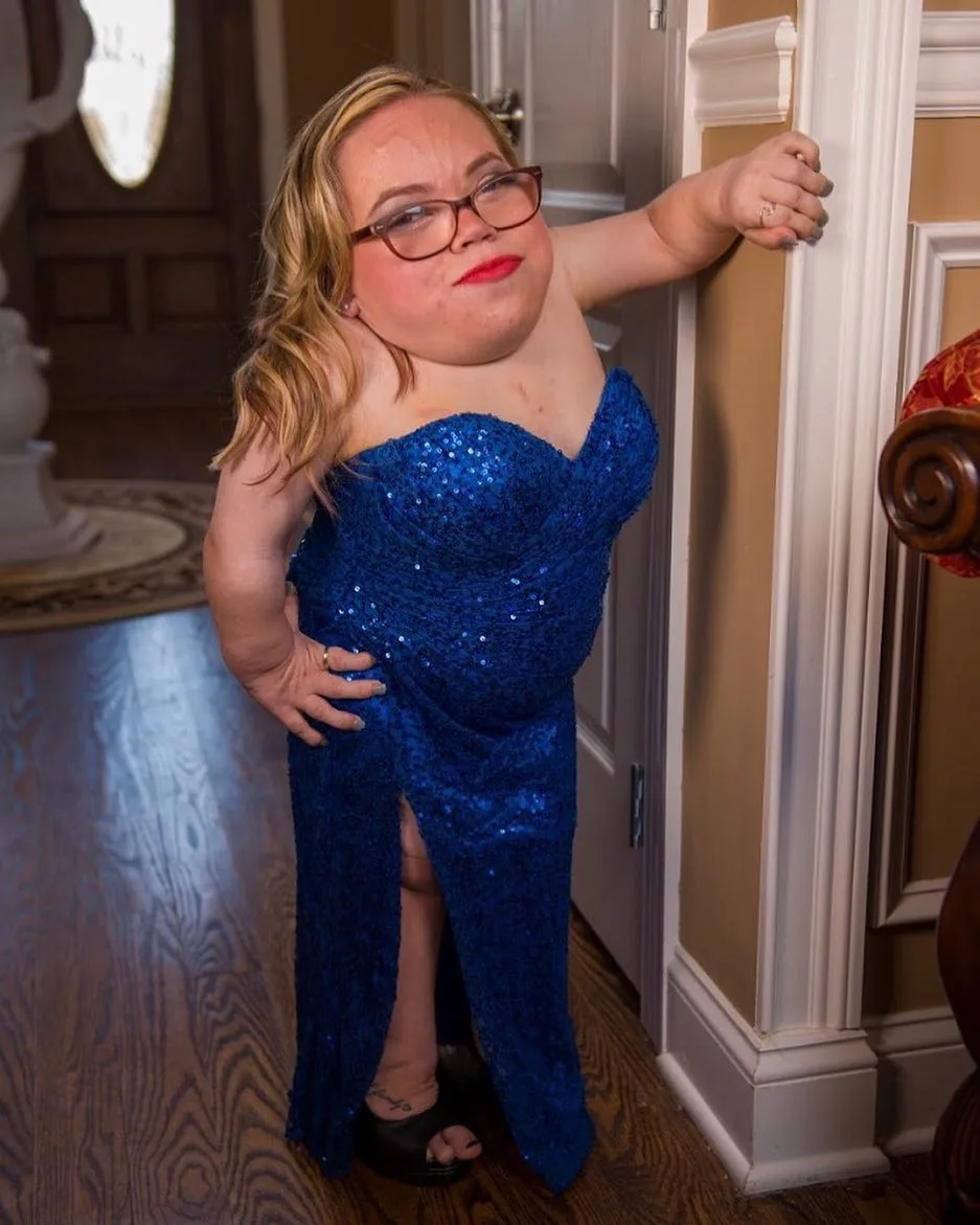 Woman with blonde hair wearing glasses and blue sparkly dress, posing next to door.