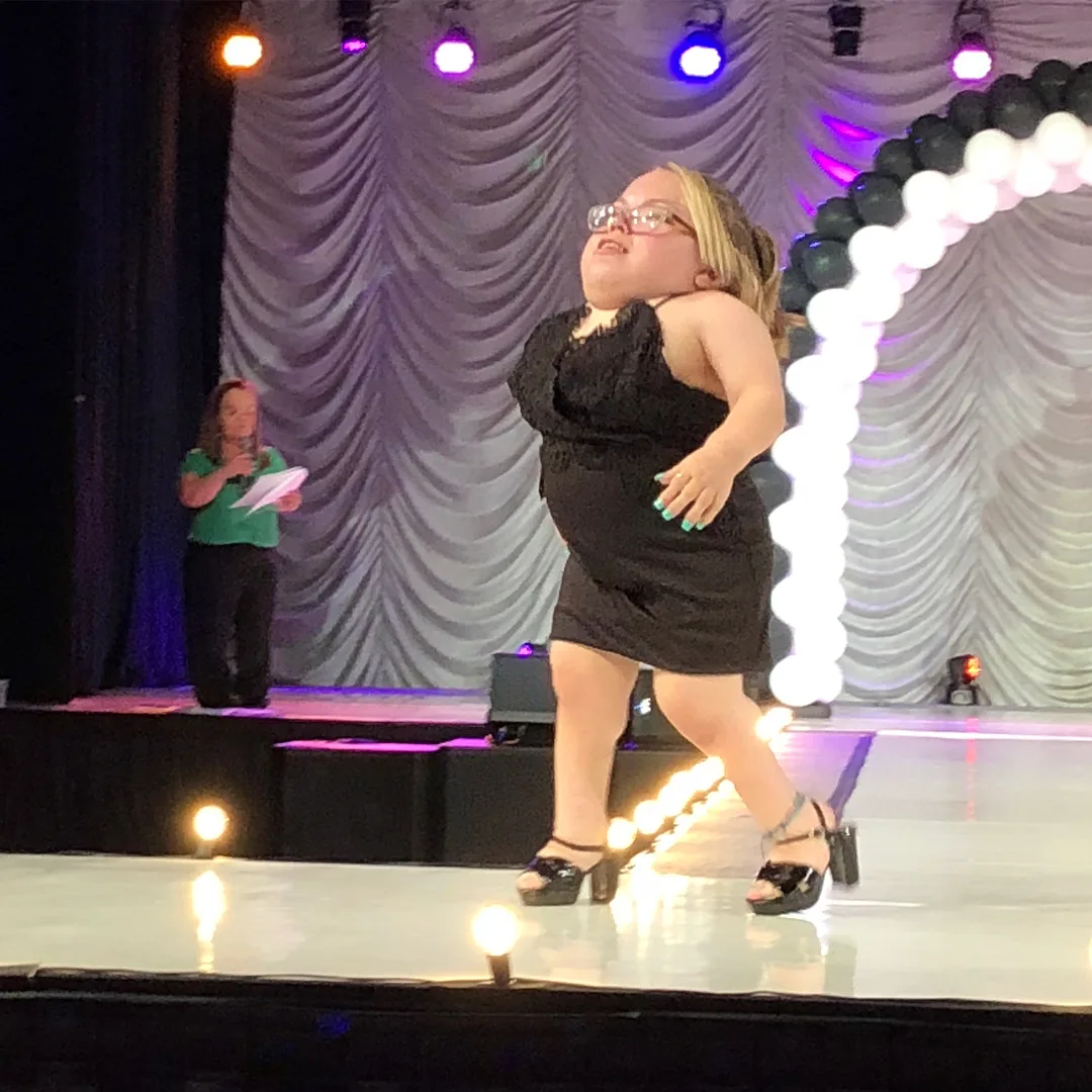 Woman with dwarfism wearing short black dress and black heels, walking down a white runway.