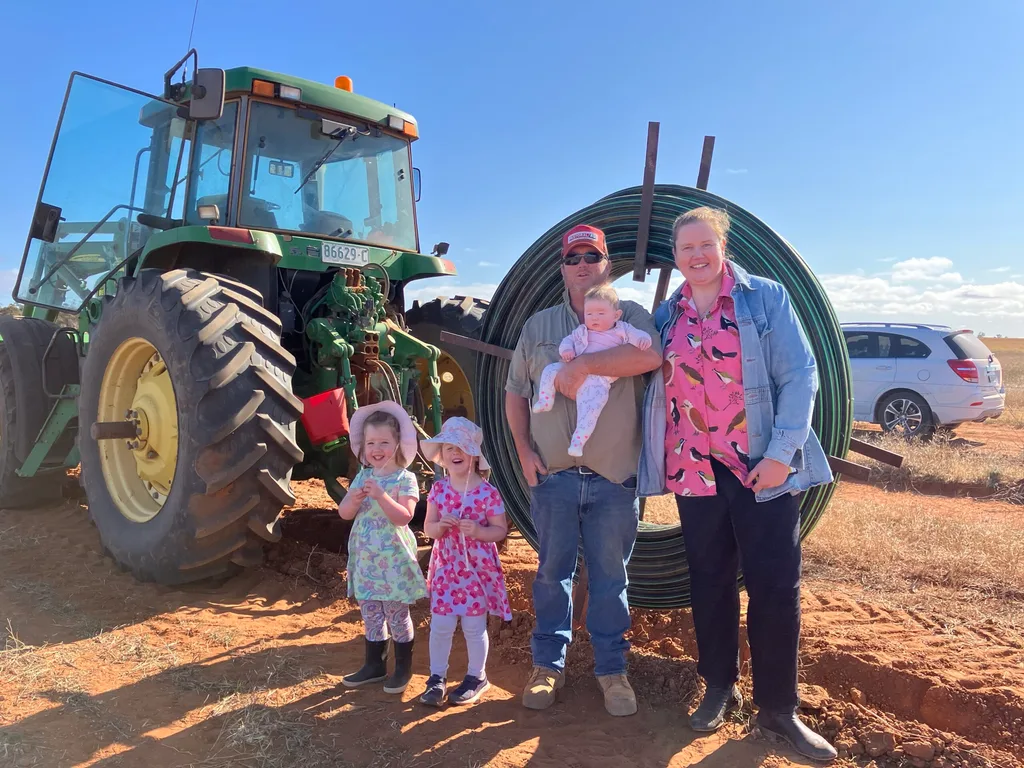 Woman in black jeans andpink shirt and man in red hat and jeans black trousers and jeans and denim jackets with two children in dreses and man holding baby in front of tractor